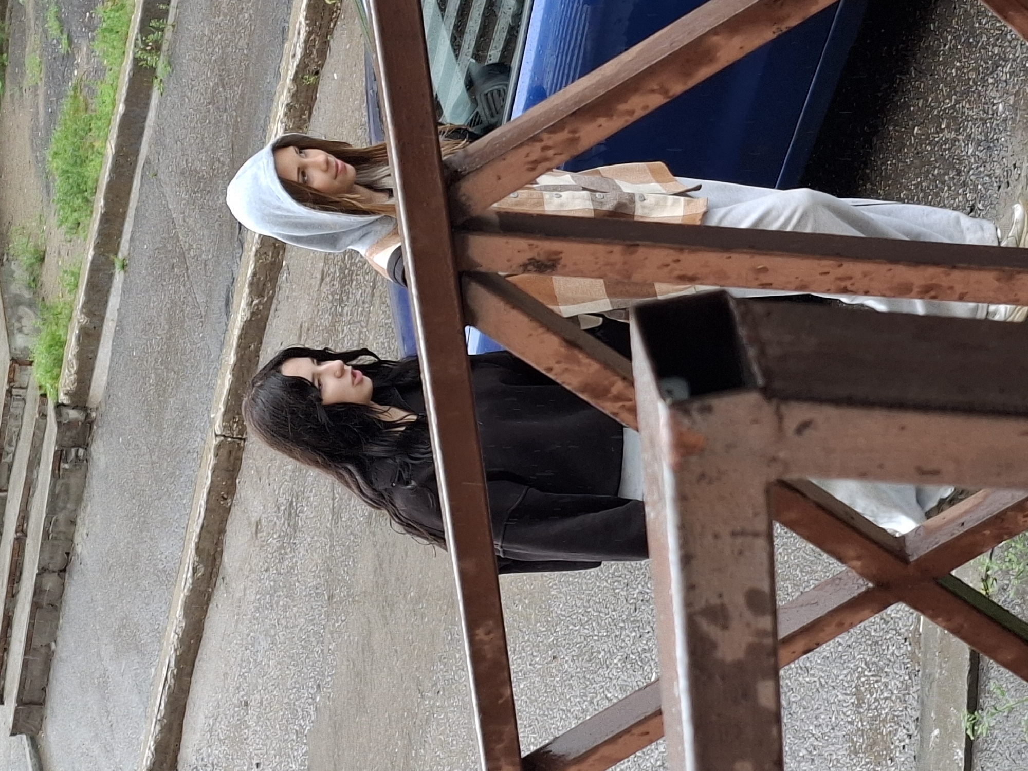 Two young females are walking down an outdoor concrete stairway. The female on the left has long, dark, wavy hair and is wearing a black hooded sweatshirt and light-colored pants. The female on the right has light brown hair and is wearing a hooded gray sweatshirt under a plaid shirt and light-colored pants. A blue car is visible parked next to the stairway, and a rusty metal railing is in the foreground, obscuring part of the scene. The ground is wet, indicating it may be raining or has recently rained. The environment appears to be an urban setting.