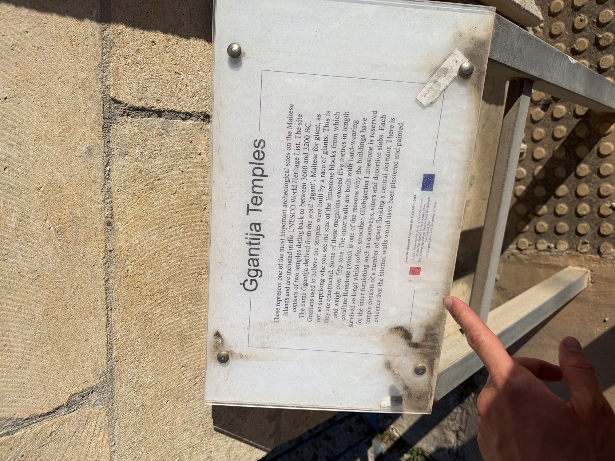 A signpost with information about the Ggantija Temples is displayed outdoors. The sign is made of a white material and is covered in dirt and smudges. A person's hand is visible pointing at the sign. The background consists of a stone wall and a metal stand supporting the sign. The stone wall is made of large, rough-hewn blocks. The ground is covered in a pattern of circular indentations. The scene appears to be in Xagħra, Malta, as indicated by the sign's text which references the Ggantija Temples.