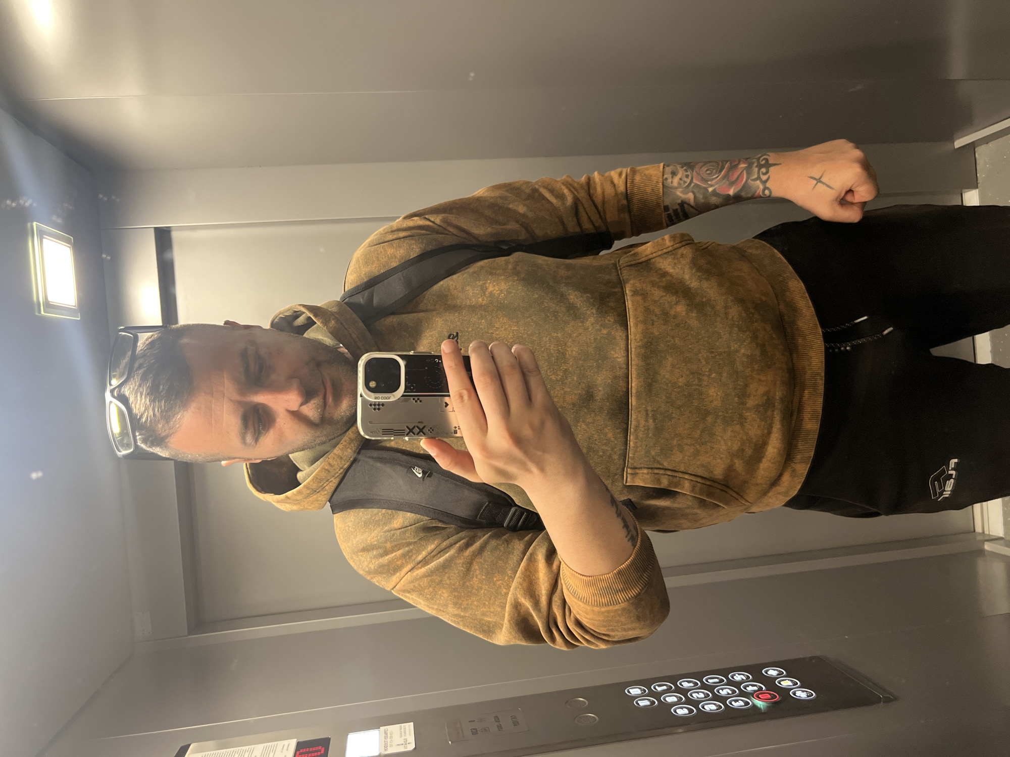 A man is taking a mirror selfie inside what appears to be an elevator. He is of light complexion, with short dark hair, stubble, and sunglasses resting on top of his head. He wears a brown and gold distressed-look hooded sweatshirt over a black t-shirt, and dark pants or shorts. A backpack strap is visible across his chest, bearing a "NIKE" logo. His left hand holds a smartphone with a silver/clear case, displaying partial text like "20 COOR" and "EXX" on its back. Tattoos are visible on both forearms, including a prominent rose and clock design, abstract patterns, and two X-like symbols on the back of his right hand, which is clenched into a fist.
The elevator interior features grey walls and floor, with reflective surfaces. An illuminated rectangular light fixture is visible on the ceiling. A control panel with numerous buttons is located on the lower right, featuring numbered buttons (0 through 32), door open/close icons, and an alarm bell symbol. A single red button, possibly an emergency stop or alarm, is distinctly illuminated. Small, difficult-to-read text on adjacent panels includes the partially visible phrase "IMPORTANT EQUIMP". The man's black pants or shorts have a white logo or text that reads "TRAIL" on the left thigh. The overall setting suggests an indoor urban environment, likely in Varna, Bulgaria, as provided.