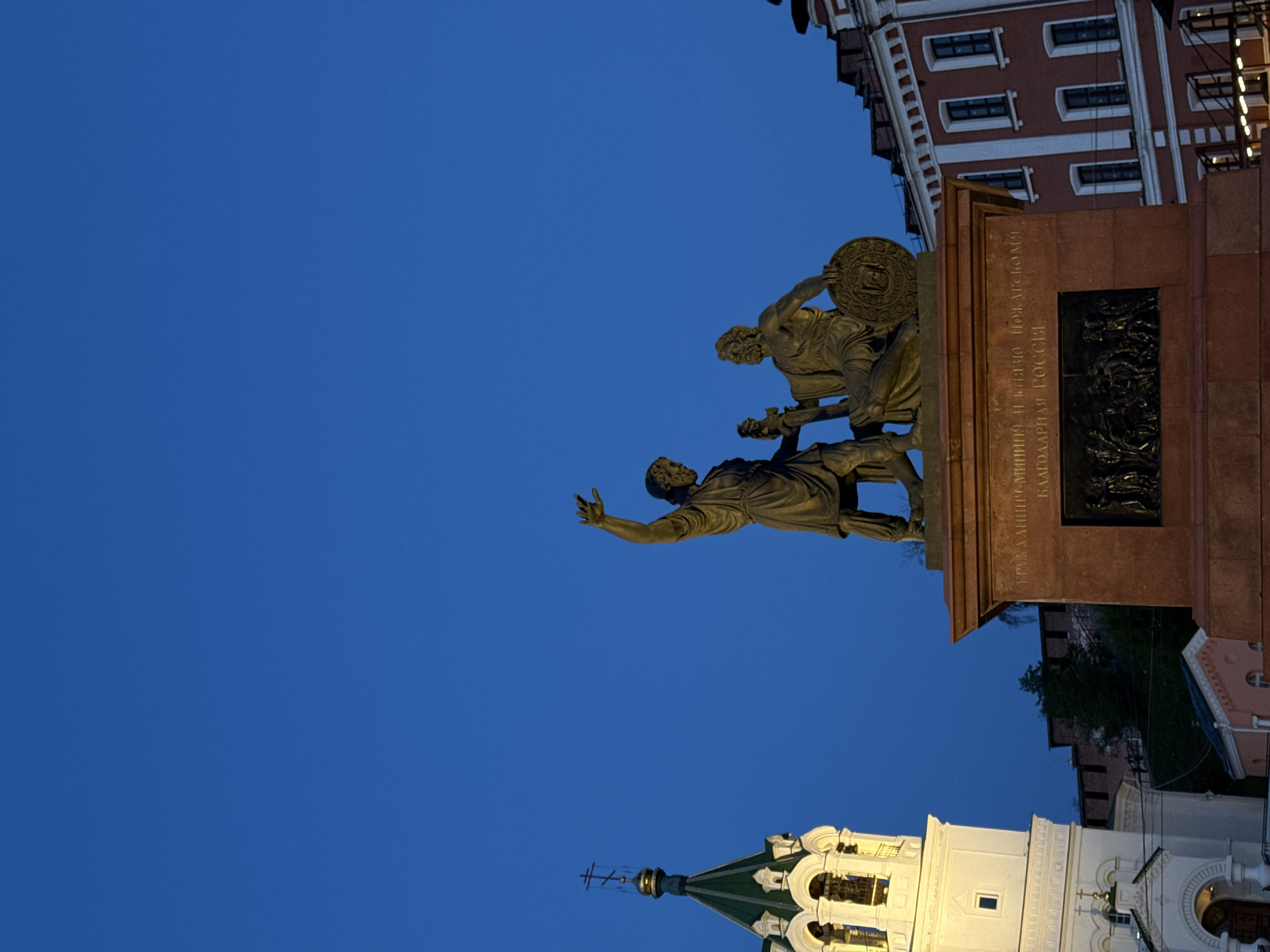 The image captures a statue in Nizhny Novgorod, Russia, against a blue sky. The statue depicts Kuzma Minin and Dmitry Pozharsky, figures known for leading the Russian resistance against Polish invaders in the early 17th century. The statue rests on a pedestal inscribed with Cyrillic text. In the foreground, there's a portion of an orthodox church with a green rooftop and white walls. A corner of a brick building, likely a historical structure, is visible in the background. The perspective suggests the photo was taken from a low angle.