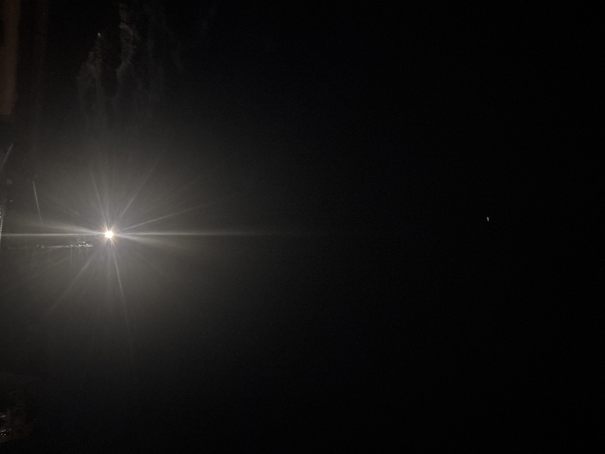 The image shows a dark night scene. A bright light source, possibly a street lamp or vehicle headlight, is located in the left portion of the frame, emitting a strong glare and starburst effect. The surrounding area appears to be shrouded in fog or mist, obscuring details. On the right side of the frame, a small, indistinct green light is faintly visible against the otherwise dark background. The specific location in the city of Lom, Bulgaria is not visually discernible.