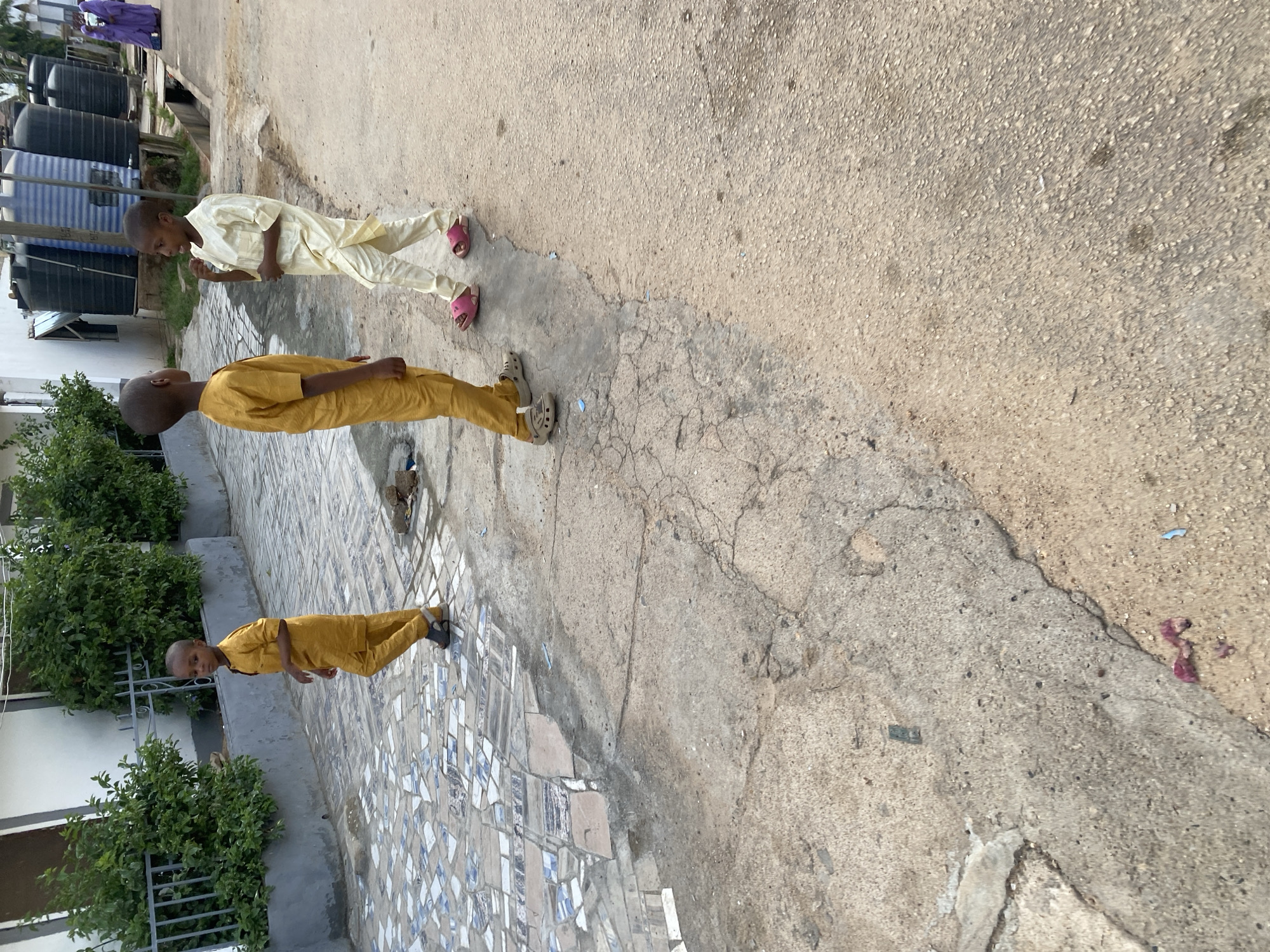 In Jos, Nigeria, a group of children walk down a street. Three young boys in yellow and cream-colored traditional clothing are prominently featured. One boy in yellow walks away from the camera on the left. In the center, another boy in yellow faces left, appearing to talk to a boy on the right dressed in cream. The boy in cream faces left, with his right hand near his mouth. They are on a cracked concrete sidewalk with a tiled section to their left. In the background, large black water tanks are visible. A woman in a purple garment walks in the distance on the right. The environment suggests an urban setting with buildings and vegetation.