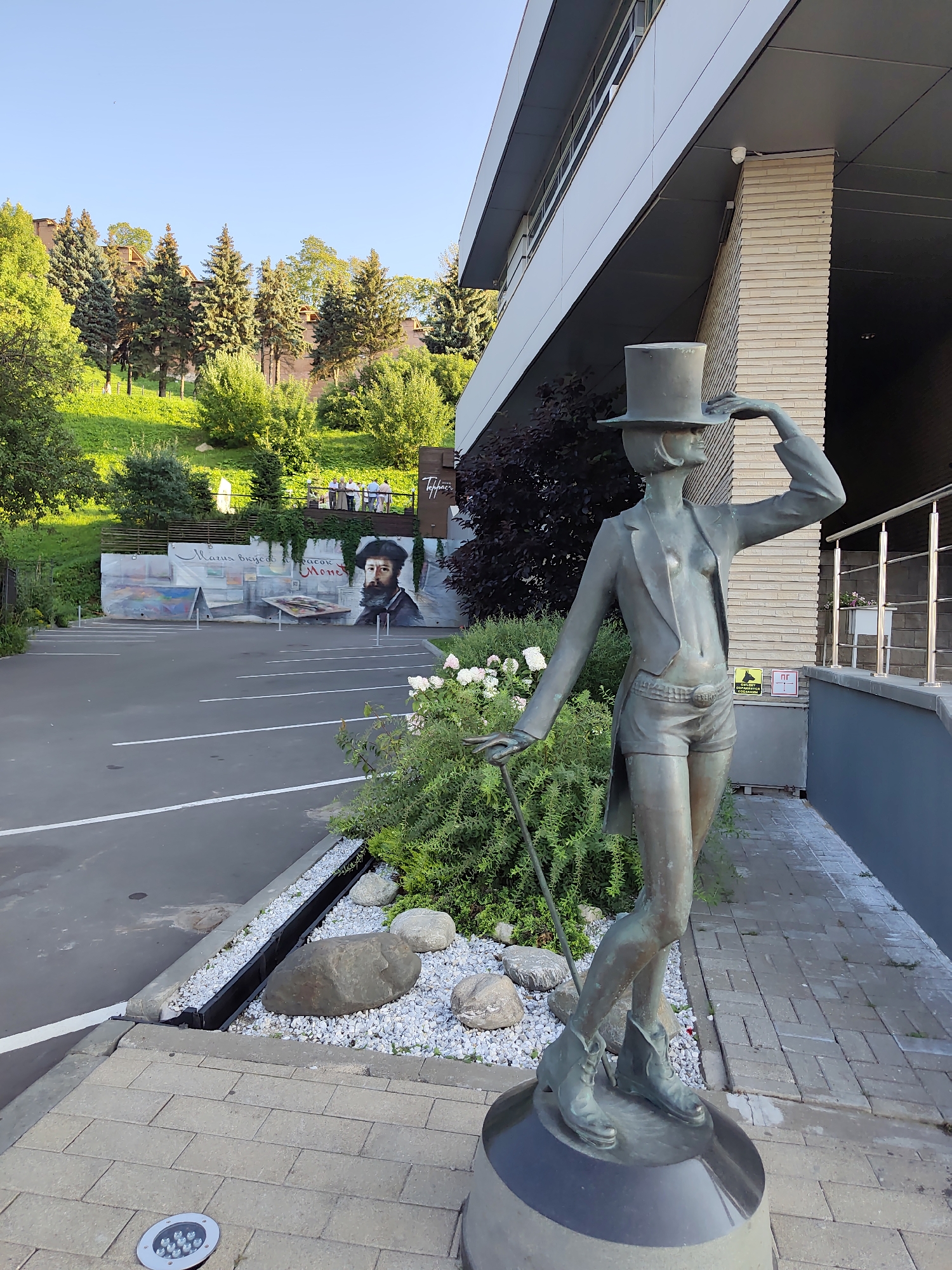The image shows a bronze statue of a woman in a top hat and a tailcoat, wearing shorts and boots, holding a cane. The statue is positioned in front of a modern building with a slanted roof and a brick facade. To the left of the statue, there is a parking lot and a hillside with trees and a mural. The mural depicts a portrait of a man with a beard and several paintings. The building appears to be a commercial establishment, possibly a restaurant or a gallery, as suggested by the sign "Tobbi" visible on the mural. The overall scene suggests an outdoor setting in a city, likely in a pleasant climate given the greenery and clear sky. The mural and the style of the building suggest a location in a European city. Further investigation reveals that the mural is a reproduction of a painting by Monet, and the statue is likely located in Nizhny Novgorod, Russia, specifically near the Verkhny Volga Embankment, an area known for its cultural attractions and scenic views.