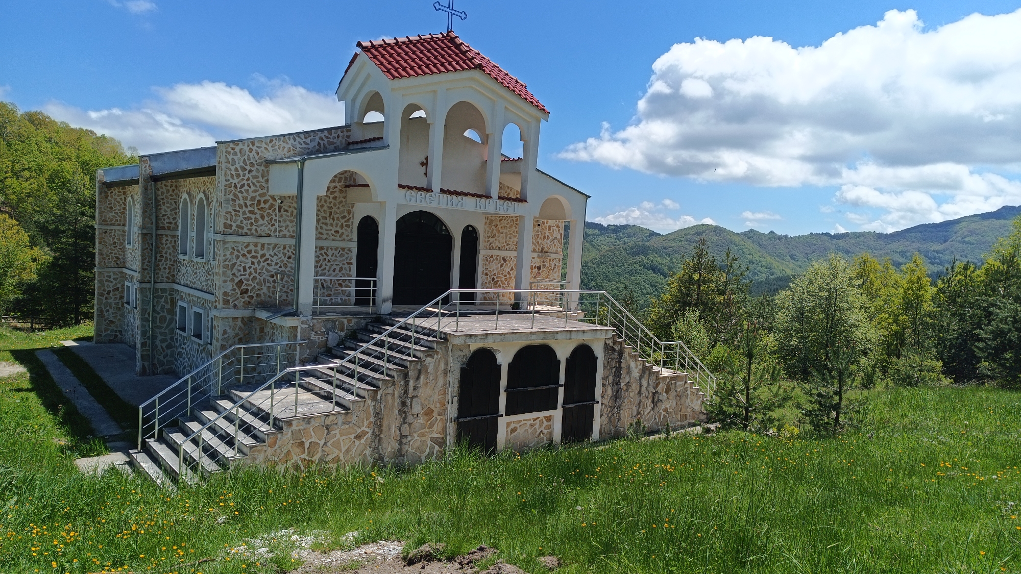 The image depicts a stone-and-white building with a red-tiled roof, identified as the Holy Cross Church located in the city of Sini Vrah, Bulgaria. The church has multiple levels, with stairs leading to the main entrance. The entrance is marked with an inscription in Bulgarian. The building is surrounded by a grassy area dotted with yellow wildflowers. In the background, there are forested hills under a blue sky with scattered clouds. No people are visible in the scene.