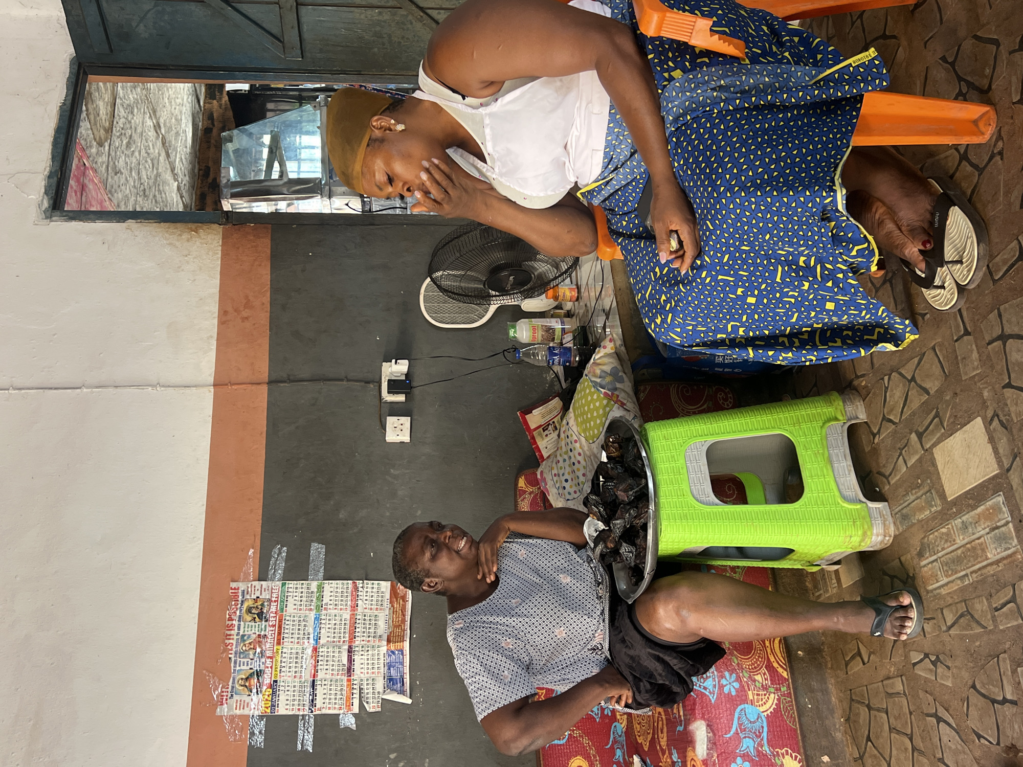 In Nkpor, Nigeria, two women are seated in what appears to be an open-air market stall or a shaded outdoor seating area. The woman on the left, with short gray hair, is wearing a blue and white patterned blouse and a black skirt. She is leaning back on a colorful patterned cushion, with her right hand resting on her chin and her left arm on her lap. In front of her, on a bright green plastic stool, is a metal tray filled with dark, dried food items, possibly fish or meat.
The woman on the right is wearing a white tank top over a colorful blue and yellow patterned skirt, with a brown headscarf. She is seated in an orange plastic chair and has her hand raised to her mouth, appearing to be speaking or perhaps holding something to her nose. To her left, on a table, is a black oscillating fan and several bottles, including what looks like water and possibly medicinal products.
The background wall is painted in horizontal bands of dark gray and orange, with calendars and posters taped to the upper section. Electrical outlets are visible on the dark gray section of the wall. The floor is tiled with a pattern of brown and beige squares. A doorway with a metal frame and a blue door is visible to the right, leading into a darker interior space. The overall impression is of a busy, lived-in environment typical of a market setting.