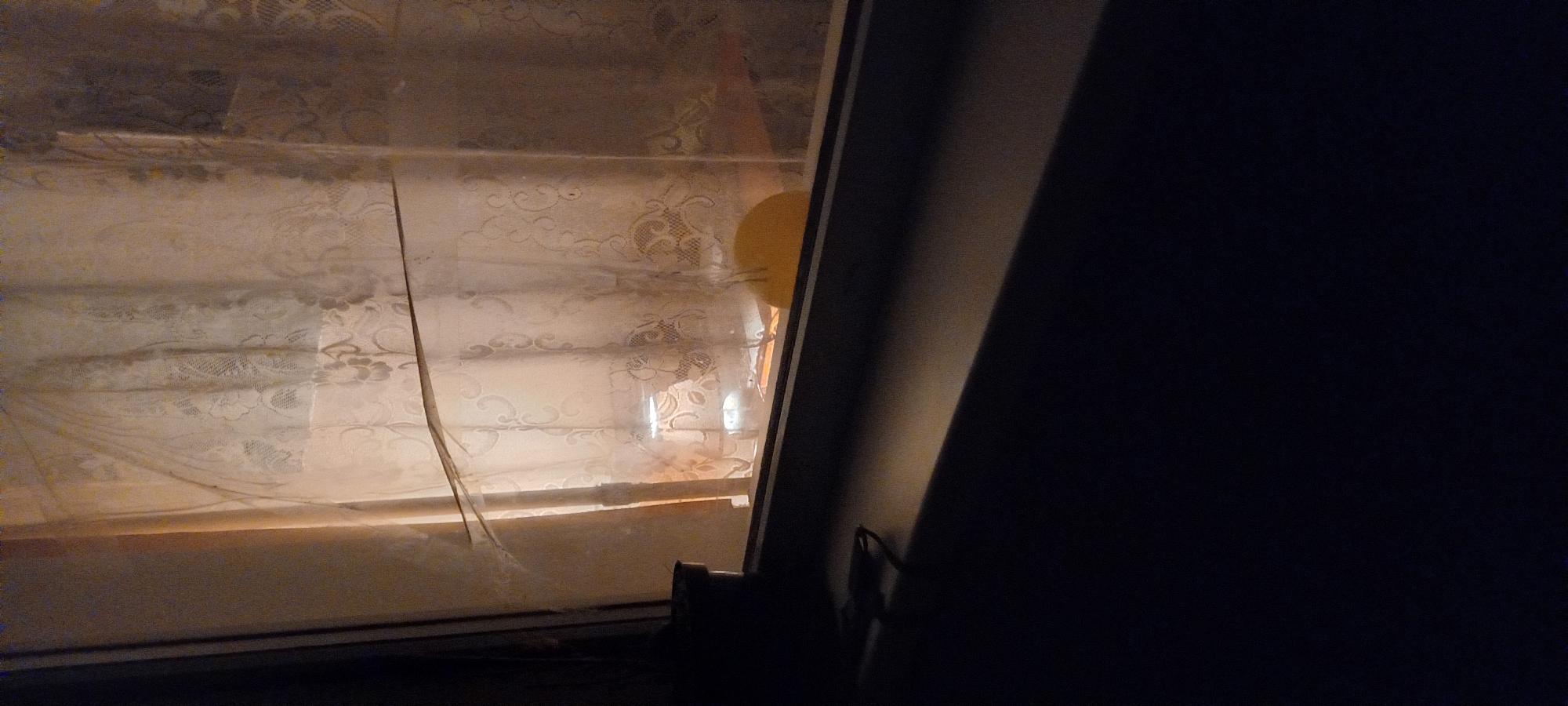 A close-up, eye-level view looks through a window with sheer, patterned curtains. The curtains are slightly torn and pulled back, revealing a dimly lit interior. A small, white lamp with a distinct shade is visible, casting a warm glow on what appears to be a surface beneath it, possibly a desk or shelf. A yellow, circular object, perhaps a plate or a toy, is also present in the background. A dark potted plant sits on the left side of the windowsill. The exterior view is not clear due to the low light conditions and the curtains. The image does not provide any specific details that would allow for identification of its location in Kryvyi Rih, Ukraine.