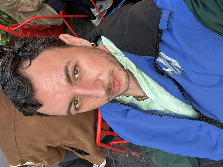 A male individual with dark, curly hair, dark eyes, and a short beard faces the camera with a neutral expression in a medium close-up shot. He wears a light mint green collared shirt under a partially unzipped blue, dark green, and black paneled jacket, revealing a silver chain necklace. His right ear displays a small stud and a larger gold-toned hoop earring.
The subject is seated in an outdoor setting, flanked by two other individuals whose backs are partially visible. To his left, a person in a brown jacket sits on a red metal chair. To his right, another person in a brown plaid jacket with elbow patches and grey sneakers sits on a similar red metal chair. Red metal chair frames are visible throughout the background. The ground is a paved surface, and some green foliage is discernible in the upper background. This scene, characterized by the distinctive red metal chairs, is consistent with an outdoor café or public seating area in Paris, France.