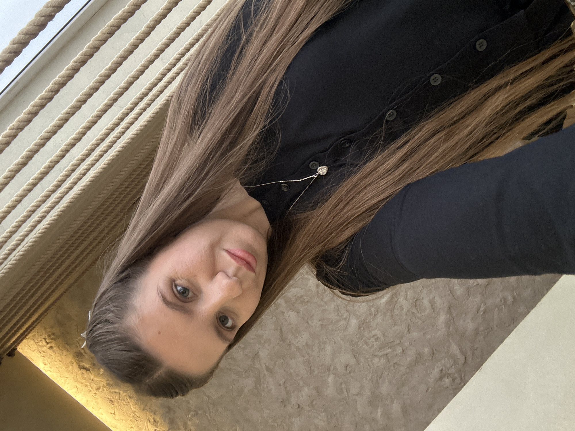 A woman with long, straight, light brown hair is the central subject of this image, captured from a low, upward-facing angle. She is positioned diagonally, with her head in the lower left quadrant and her body extending towards the upper right. She wears a dark, long-sleeved top, possibly black or navy, featuring several buttons down the front. A delicate silver chain necklace with a small, heart-shaped pendant adorns her chest. Her face is turned directly towards the camera, showing light-colored eyes and a subtle, neutral expression with a hint of a smile.
The background consists of several elements. In the upper left, multiple parallel beige ropes or cords extend diagonally across a lighter surface, suggesting a decorative or structural element like a railing or window treatment. The right and lower portions of the background reveal a textured wall with an uneven, plaster-like finish. A warm, golden light source or reflection is visible in the lower left, near her head. The lighting appears soft and natural for an indoor setting during the daytime. No text is visible. The scene is set in Klin, Russia.