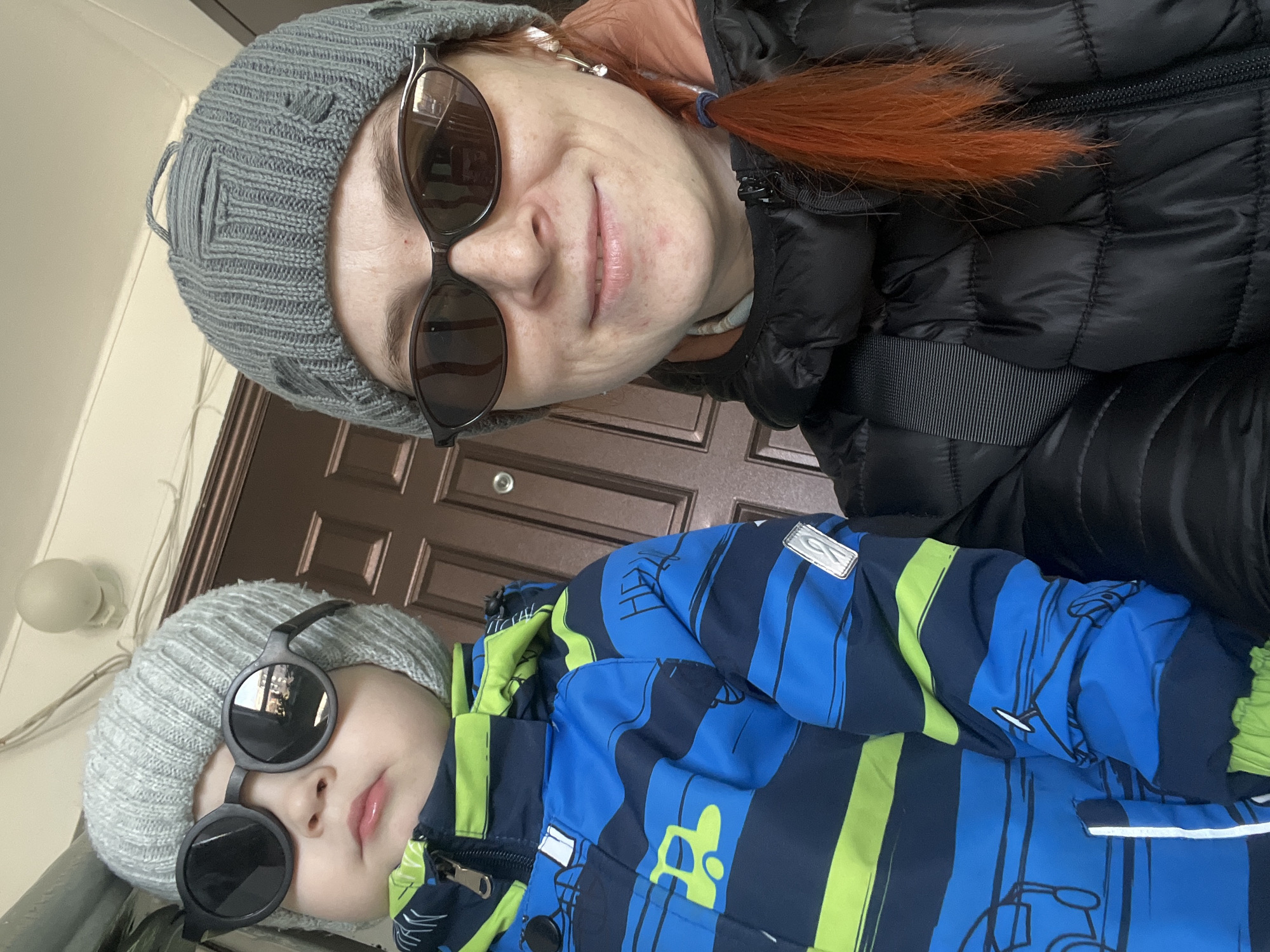 A selfie features an adult and a child, both wearing sunglasses and hats. The adult has reddish-orange hair pulled back into a ponytail. The child is wearing a blue and green striped jacket. A brown door is visible in the background. The location is Chaikovsky, Russia.