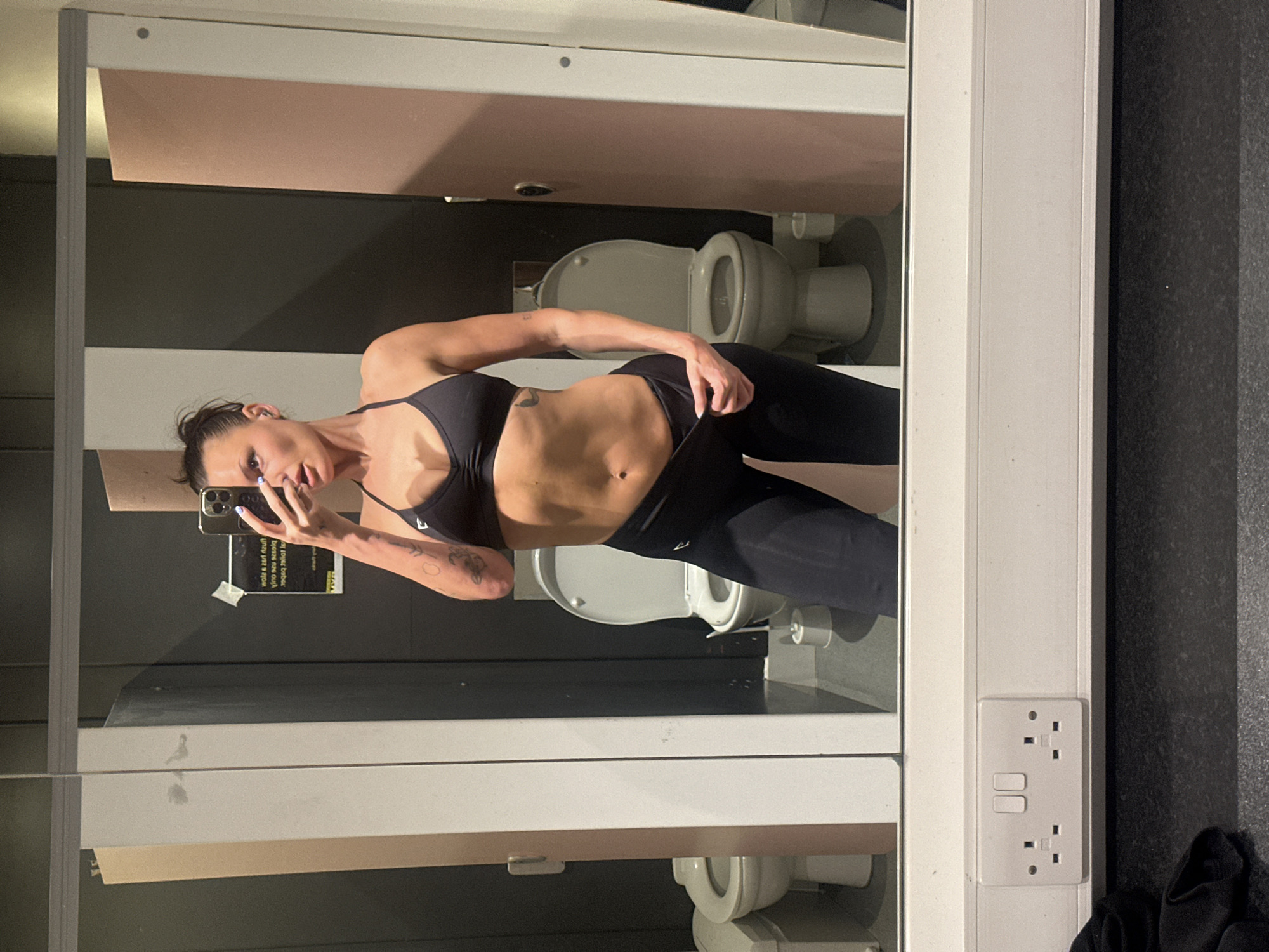 A woman in a black sports bra and leggings takes a selfie in front of a mirror. The reflection reveals a public restroom setting with multiple toilets and toilet paper dispensers. A British-style electrical outlet is visible on the right side of the mirror frame. The exact location in Tunbridge Wells cannot be determined.