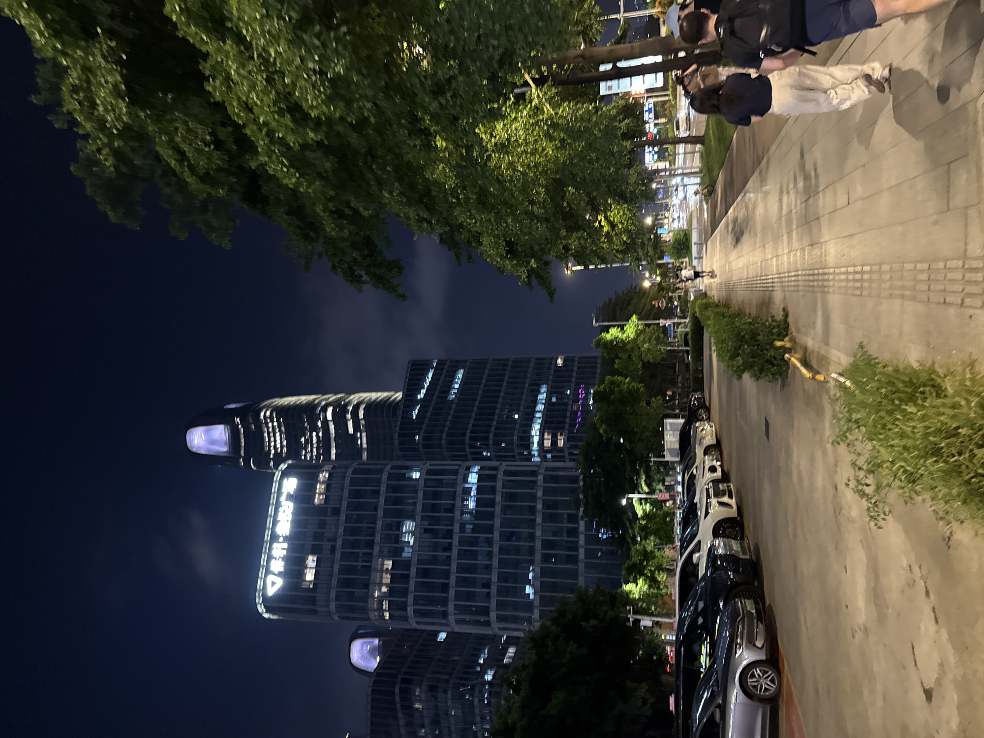 In the city of Haizhu District, China, a nighttime scene unfolds on a wide, paved sidewalk adjacent to a parking lot filled with cars. Tall, modern buildings with illuminated windows dominate the background, one of which features a distinctive glowing top. Trees line the side of the sidewalk, their leaves lit by streetlights. Several people are walking on the sidewalk, including a couple in the foreground with the male wearing a backpack. Further down the sidewalk, other individuals are visible. The parking lot contains a variety of vehicles, including sedans and vans, parked in neat rows. The overall atmosphere is urban and modern, with the artificial lights of the city illuminating the scene against the dark night sky.