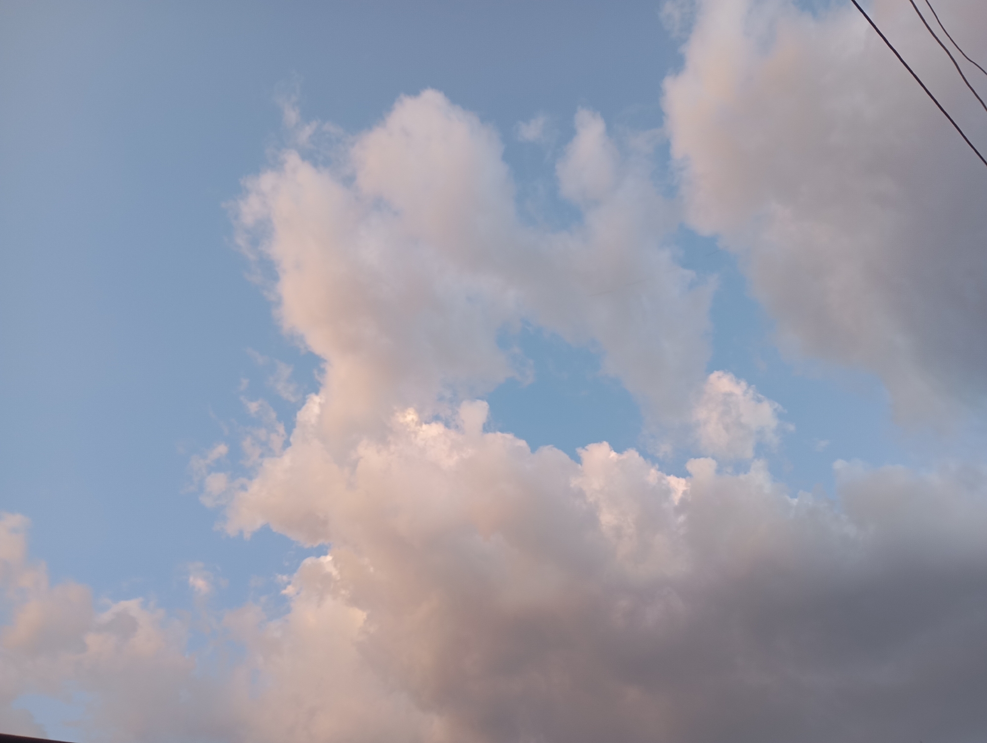 The image shows a sky with clouds. The clouds are white and pinkish, and the sky is blue. There are also some power lines visible in the upper right corner of the image. The image appears to have been taken during the daytime, possibly in the late afternoon or early evening, given the warm tones of the clouds. There are no people or any other discernible objects or activities in the image. The scene is natural and serene.