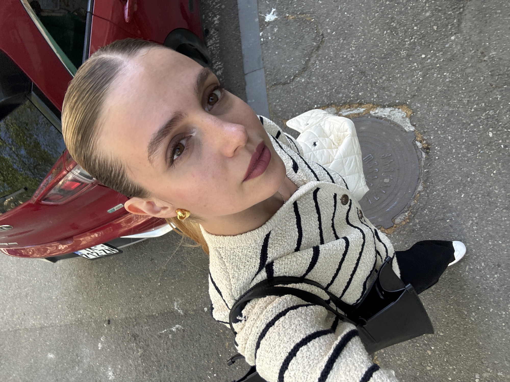 A high-angle selfie depicts a young woman looking directly upwards at the camera in an outdoor urban setting in Tbilisi, Georgia. She has light skin, brown eyes, and her light brown hair is neatly styled and slicked back with a visible side parting. Her expression is neutral to slightly serious, with dark lipstick applied to her lips. She is wearing a cream-colored knit sweater with thick black horizontal stripes, and a small black shoulder bag is visible on her left side. A small, gold-colored, knot-shaped earring adorns her left ear. A white quilted jacket or vest is draped over her left arm or shoulder.
The background reveals a paved asphalt surface, suggesting a street or sidewalk. To the left, the rear quarter panel and bumper of a red car are visible, with its license plate clearly displaying "232-ZN" and "GEORGIA" written above it. On the ground to the right of the woman, a circular metal manhole cover is partially seen, surrounded by cracked asphalt. The lighting indicates a clear, bright daytime setting.