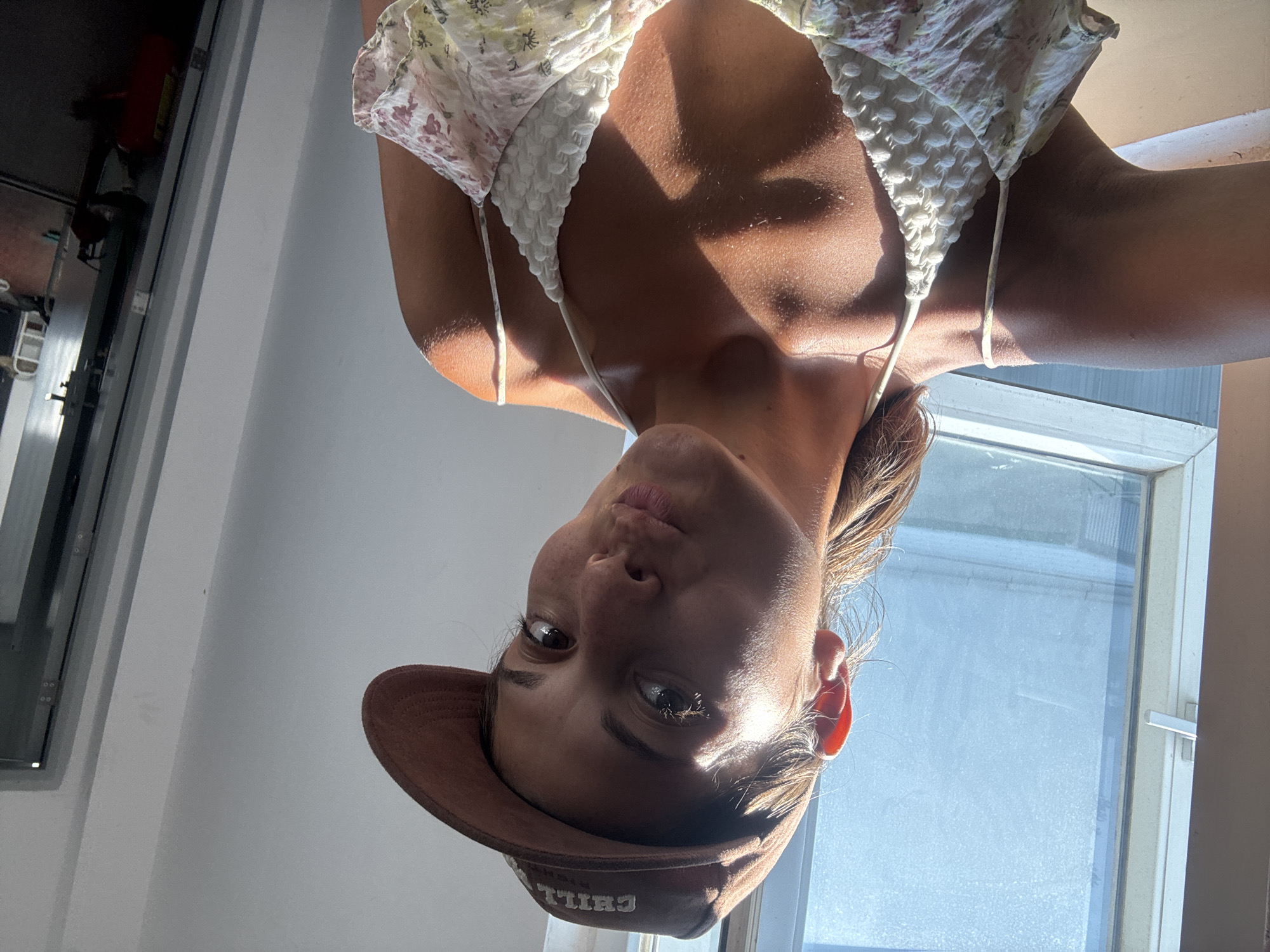 A young woman wearing a floral patterned top and a brown baseball cap with the word "CHILL" stitched on the front is positioned in front of a window. The image is upside down. The setting appears to be indoors. Đà Nẵng, Vietnam cannot be ascertained from the provided image.
