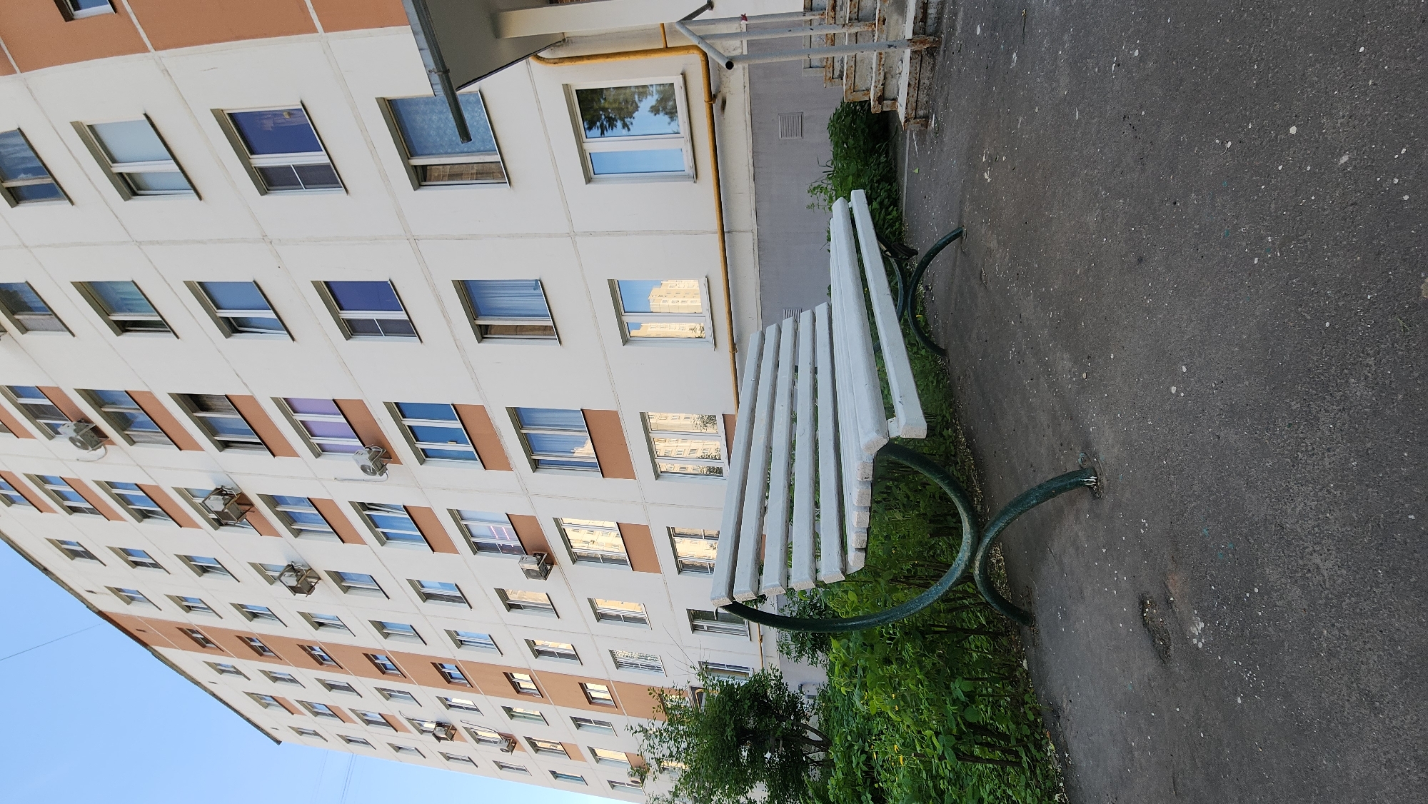 The image depicts a street scene in what is likely Moscow, Russia, based on the architectural style of the apartment building in the background. The building is multi-storied with a white facade and horizontal bands of orange/brown trim around the windows. Multiple windows are visible across its facade. An older white bench with green, circular iron supports stands on a concrete pavement. Patches of green shrubbery surround the bench and base of the building. A metallic staircase can also be seen in the background. The lighting indicates it is likely daytime. The view is shot from a low angle, looking upward toward the building.