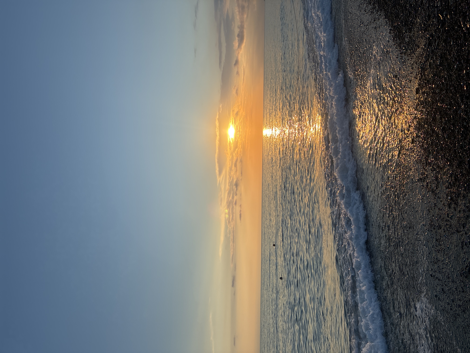 The image depicts a serene beach scene at sunrise or sunset, likely in Sochi, Russia. The sun, a brilliant orb of light, is positioned low on the horizon, casting a warm, golden glow across the sky and reflecting shimmeringly on the water's surface. Wispy clouds, tinged with orange and pink hues, are scattered across the pale blue sky.
The sea is calm, with gentle waves lapping at the shore. The water exhibits a beautiful interplay of light and shadow, with the sunlight creating a dazzling path across its surface. In the foreground, a dark, pebbly beach stretches out, wet from the receding tide and catching the last or first rays of the sun. Two small, dark objects, possibly buoys or distant birds, are visible in the water.
The overall mood is one of tranquility and peacefulness, evoking the calming beauty of a coastal sunrise or sunset. The clear, uncluttered composition emphasizes the natural elements of sky, sea, and shore. There is no visible text in the image.