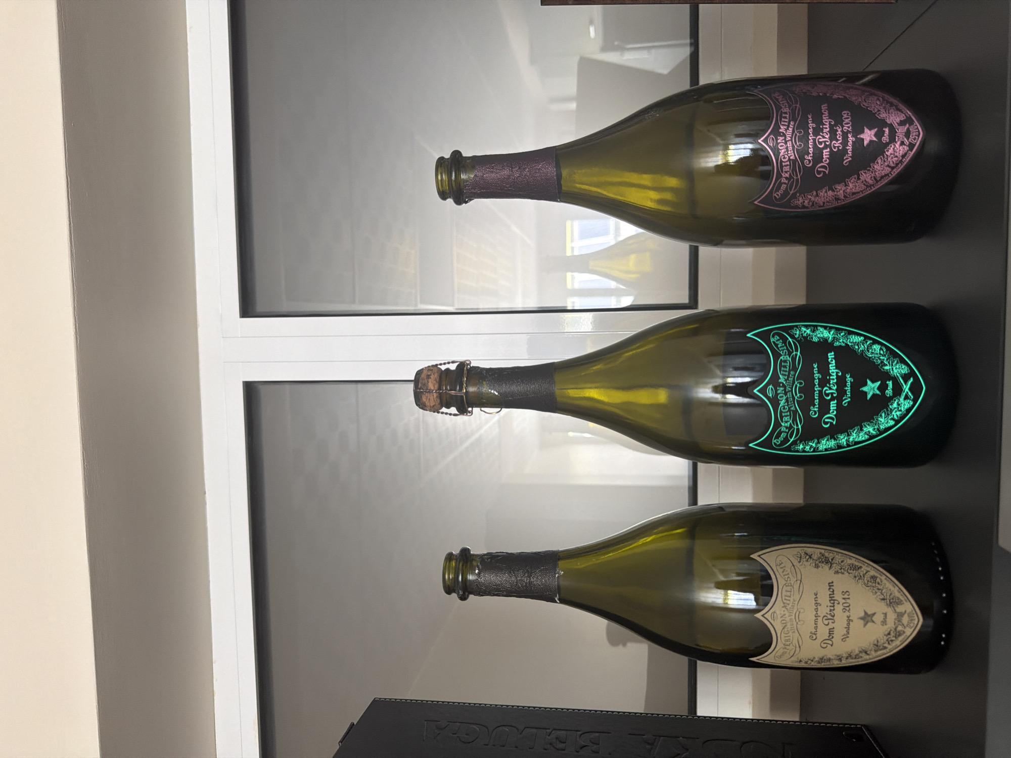 Three bottles of Dom Pérignon champagne are displayed on a dark shelf in front of a large window with reflections. The bottle on the left is Dom Pérignon Vintage 2013. The middle bottle is glowing a bright neon green and appears to be a Dom Pérignon vintage, though the year is not clearly legible. The bottle on the right is Dom Pérignon Rosé Vintage 2009. To the left of the champagne bottles is a black box with embossed text, one word of which appears to be "BELUGA." The environment suggests an indoor setting, possibly a display case or a well-lit room. There are no people or animals present in the image. The city of Dolno Ablanovo, Bulgaria is not visually identifiable from the provided image.