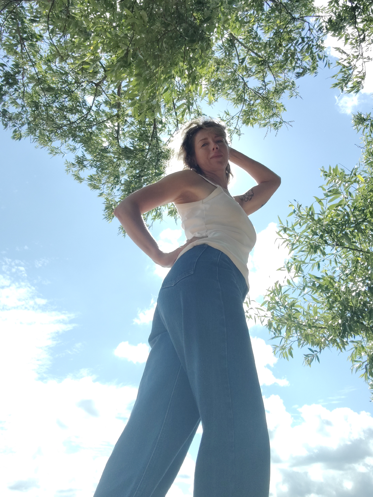 A low-angle shot captures a woman standing outdoors, looking up at the sky. She is wearing a white tank top and wide-leg blue jeans. Her left hand is on her hip, and her right hand is behind her head. Her hair is short and blonde, and she has a tattoo on her right upper arm. Sunlight streams through the leaves of a tree, creating a dappled effect. The sky is blue with scattered white clouds. The image is likely taken in an outdoor public space within Saint Petersburg, Russia, possibly a park or near a body of water, given the presence of trees and the bright daylight suggesting fair weather.