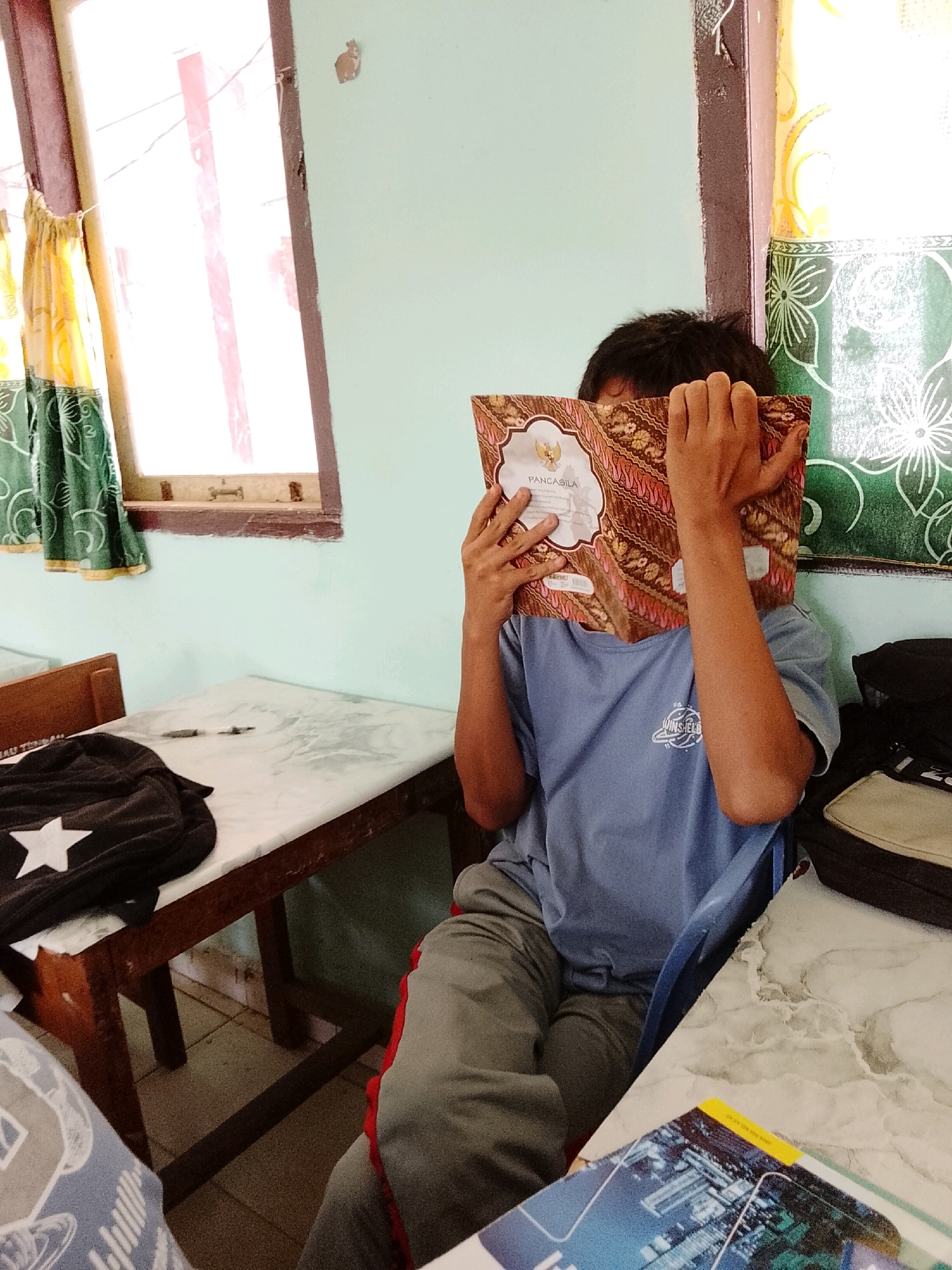 A person is sitting at a table in what appears to be a classroom or communal room in Tanjung Setia, Indonesia. The person is wearing a blue t-shirt and gray sweatpants, with their face mostly obscured by a book they are holding open. The book has a brown cover with a patterned design and the word "PANCASILA" visible. In front of the person, on the table, is a black backpack with a white star. To the left, another table is visible with a similar marble-like surface, on which a pen lies. A window with green and yellow patterned curtains is on the left side of the frame, and a light green wall is in the background. The overall impression is of a quiet, possibly indoor educational setting.