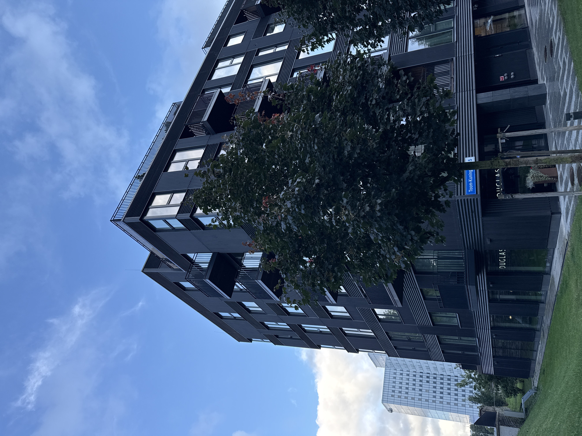 This is a street-level, low-angle, full shot of a modern, dark grey, multi-story apartment building in the city of Tallinn, Estonia. The building has numerous balconies with black railings and large windows that reflect the blue sky and scattered clouds. A mature tree with dark green foliage is partially obscuring the front of the building, and another tree is visible to the right. In the background, a tall, modern glass skyscraper with a patterned facade is visible to the left. A blue street sign that reads "Toom-Kuninga 5" is affixed to the building near the street level. The ground in front of the building is paved with patterned grey stones, and to the left is a grassy embankment. The overall scene depicts urban architecture under a partly cloudy sky.