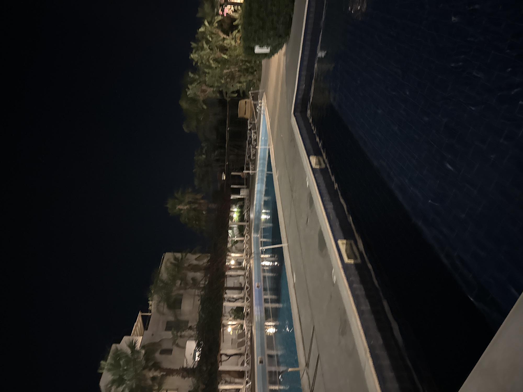 The image depicts an outdoor swimming pool area at night. The foreground shows a section of a dark blue swimming pool with a tiled bottom, partially obscured by the edge of another pool. The middle ground features a large, rectangular swimming pool filled with clear blue water, reflecting the lights from the surrounding area. A light-colored paved pathway borders the pool. Beyond the pathway, there is a covered area with lounge chairs, and behind that, a white building with multiple floors, likely a hotel or resort, surrounded by palm trees and other greenery. The sky is dark, indicating it is nighttime. The scene appears to be a resort or hotel complex in Isola delle Femmine, Italy.