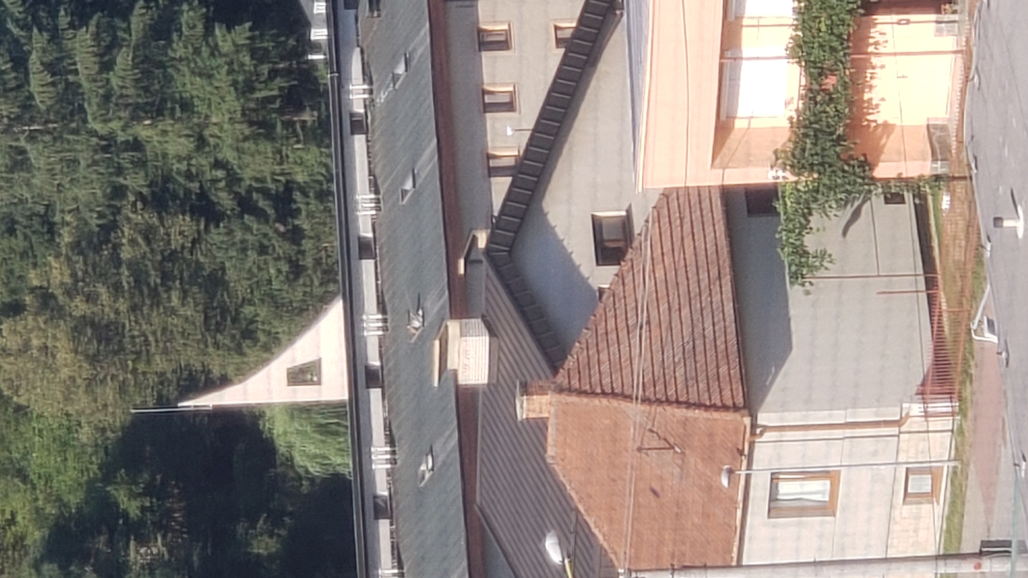 The image shows a view of buildings and trees in Prešov, Slovakia. In the foreground, there is a house with a red tiled roof. To the right of this house, there is a light-colored building with a balcony covered in greenery. Behind these buildings, there is a larger building with a dark grey roof and multiple windows. Further in the background, a triangular structure with a small window is visible, surrounded by trees. The overall scene appears to be a residential or mixed-use area.