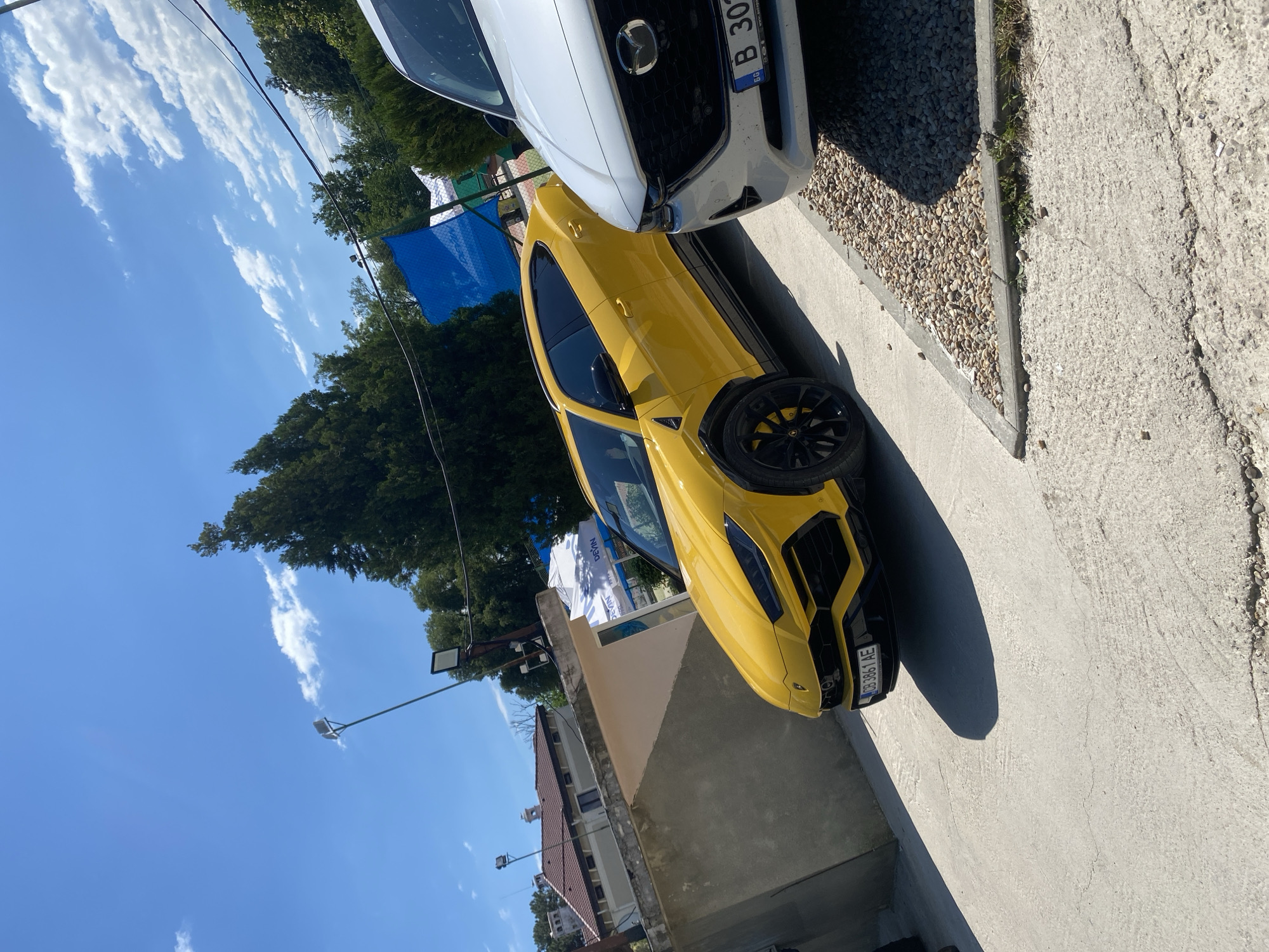 This media file depicts a bright, sunny outdoor scene featuring two cars parked side-by-side on a gravel and concrete surface. The primary subject is a vibrant yellow sports car, which is positioned in the foreground, angled towards the viewer. Its sleek design and low profile are evident, with a black wheel and yellow brake calipers visible. To its left and slightly behind is the front of a white car, identifiable by its grille and a license plate.
The setting appears to be an urban or suburban area, possibly a parking lot or a roadside. In the background, lush green trees suggest a warm-weather environment, and a clear blue sky with scattered white clouds indicates fair weather. The time of day is likely midday, given the bright sunlight and distinct shadows cast by the cars.
Visible text includes license plates on both cars. The yellow car's plate reads "CB 3861 AE". The white car's plate shows "B 30Z". There is also text on a blue banner in the background, though it is partially obscured and illegible. A street light is visible in the distance. The overall impression is one of a casual, daytime scene, possibly in Varna, Bulgaria, given the location context. No people are visible in the image.
