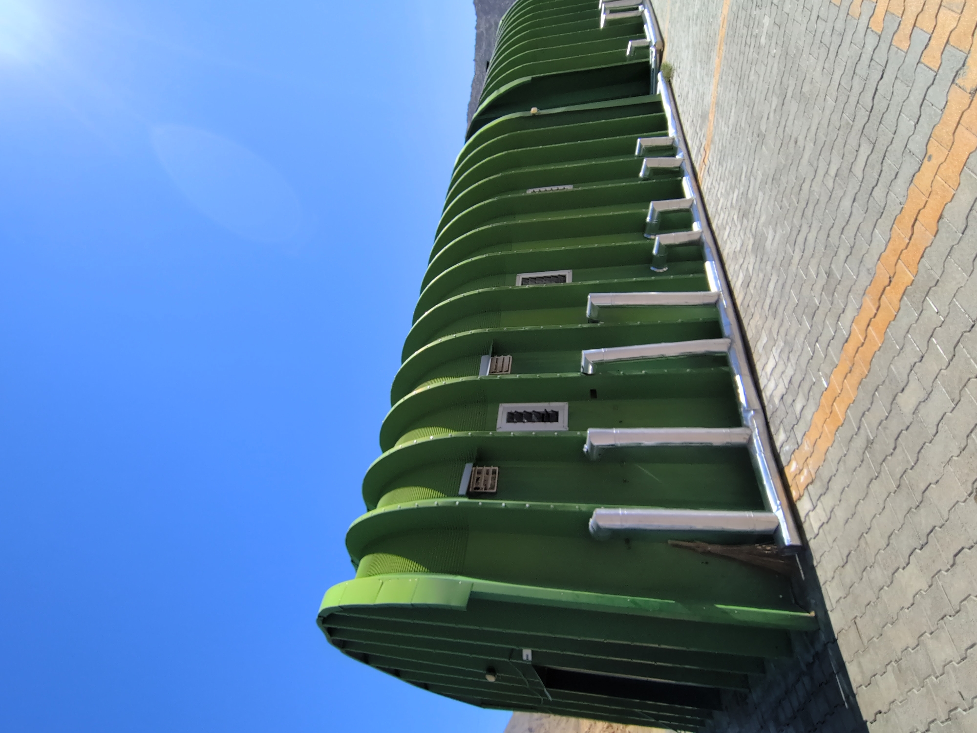 The image shows a long, arched, green metal building under a clear blue sky. The building has a ribbed structure with several rectangular openings or vents along its side. Silver-colored pipes run along the base of the building. The ground in front of the building is paved with interlocking gray bricks, with two yellow lines running parallel to each other. In the distance, a barren, mountainous landscape is visible. The scene appears to be an outdoor storage or industrial facility. There are no people or animals in the image.