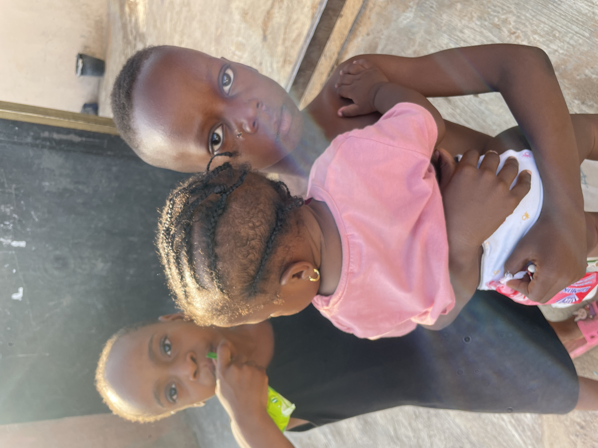 The scene shows two children and their reflections. An older child is holding a baby wearing a pink shirt and a diaper. The other child is visible through the reflection. The location is an outdoor area in Lokoja, Nigeria. The older child has a nose piercing.