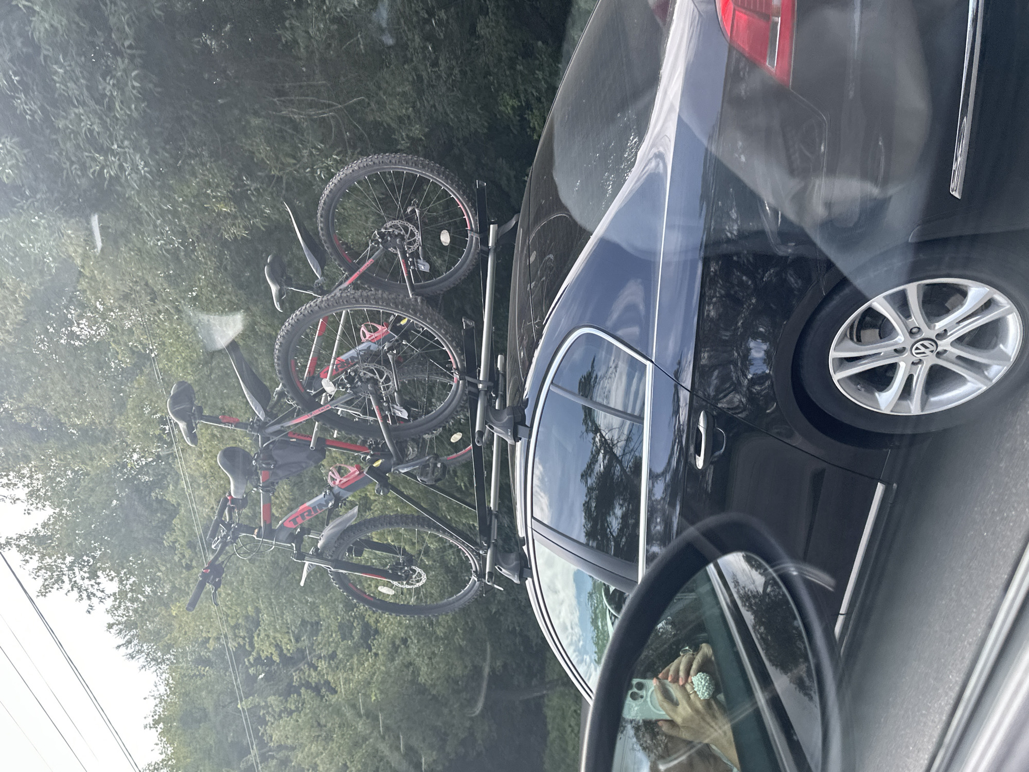 Two red and black mountain bikes are mounted on a roof rack atop a black sedan. The sedan is traveling on a road, with trees lining the background and power lines overhead. The driver's side mirror of another vehicle is visible in the foreground, reflecting a person holding a smartphone. The scene appears to be set in a suburban or rural area, given the presence of trees and the absence of tall buildings.