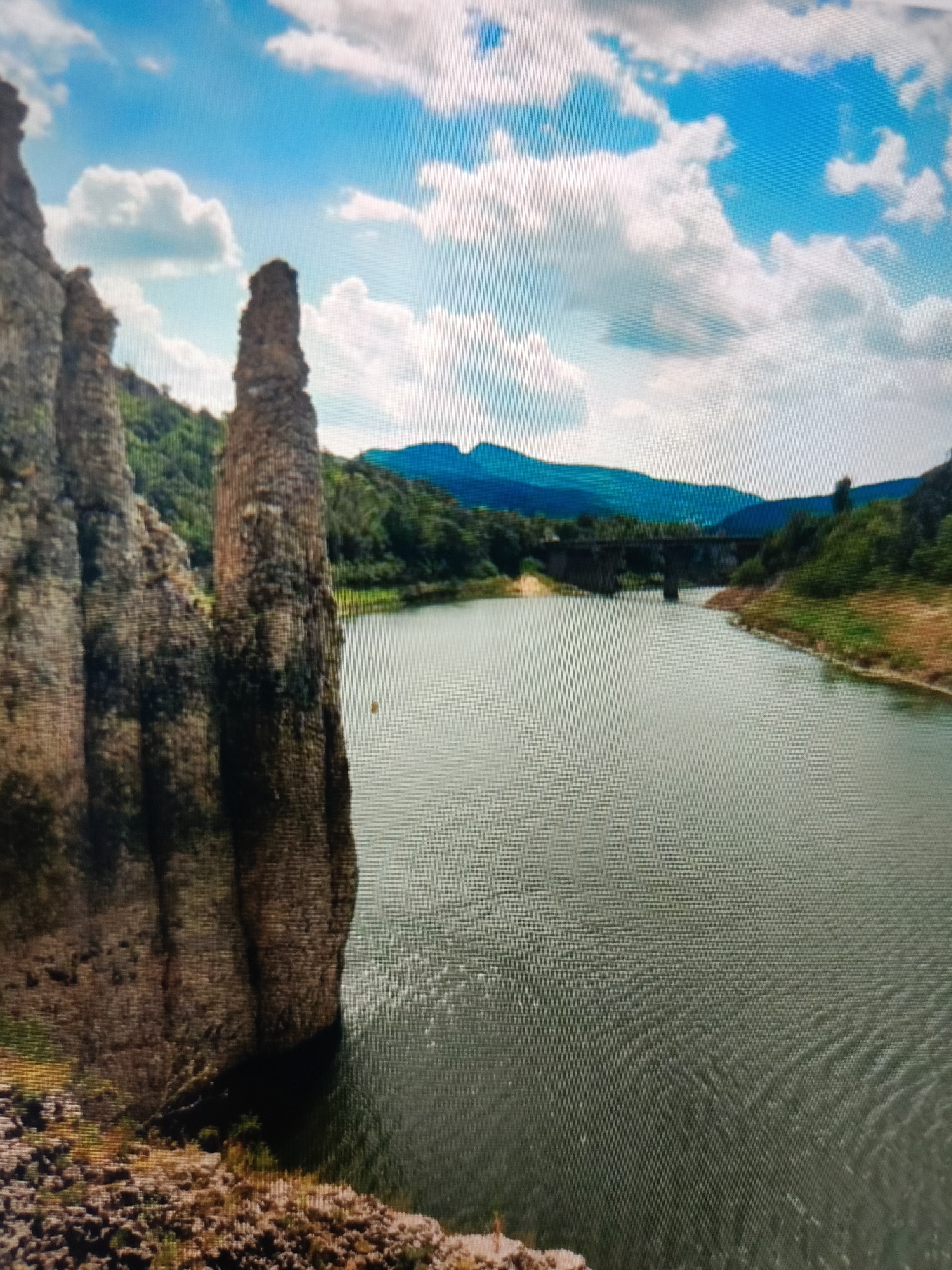 This is a photograph of a natural landscape. In the foreground, to the left, are tall, weathered rock formations with textured surfaces. Beyond these rocks, a wide body of water stretches into the distance. The water has ripples and reflects the sky. Along the far bank of the water, lush green trees and vegetation cover rolling hills. In the middle distance, a bridge with stone arches spans the water. In the background, layered mountains fade into the horizon under a bright blue sky with fluffy white clouds. The scene is devoid of people or any artificial structures other than the bridge. There is no indication that this location is in Varna, Bulgaria.