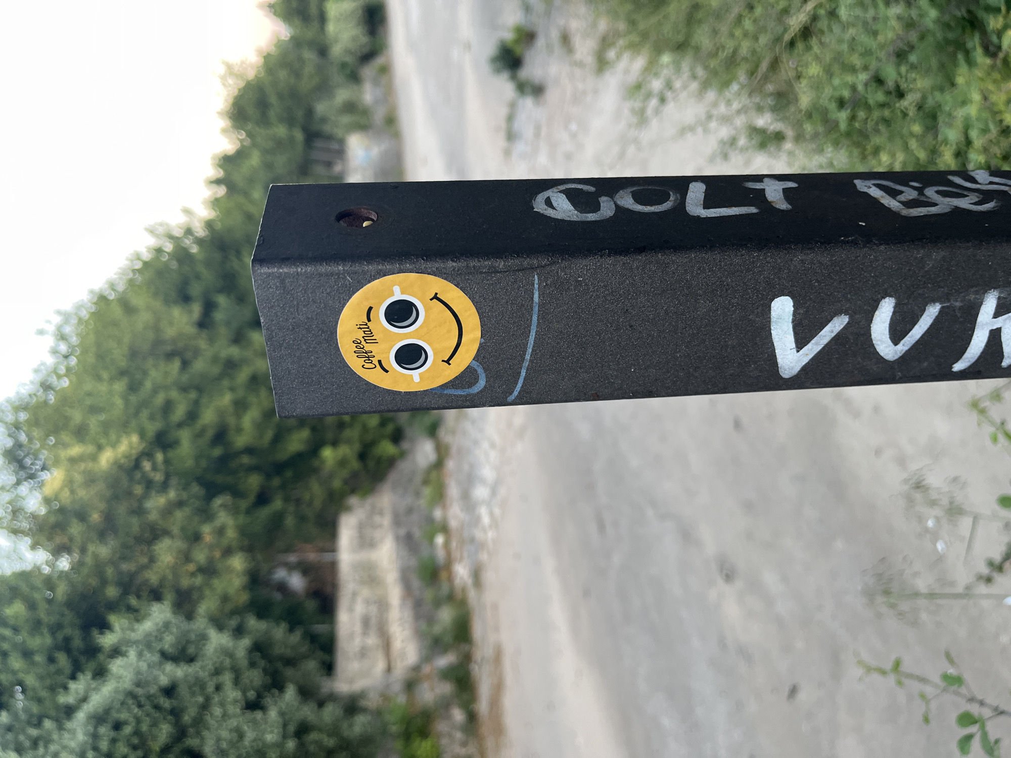 In Budva, Montenegro, a black metal post stands in an outdoor environment. The post has a round sticker with a yellow smiley face that has sunglasses and the words "Coffee Mati" printed on it. There is also graffiti on the post, with white lettering that appears to say "COLT BI" and "LUH". The background consists of trees and a dirt path.