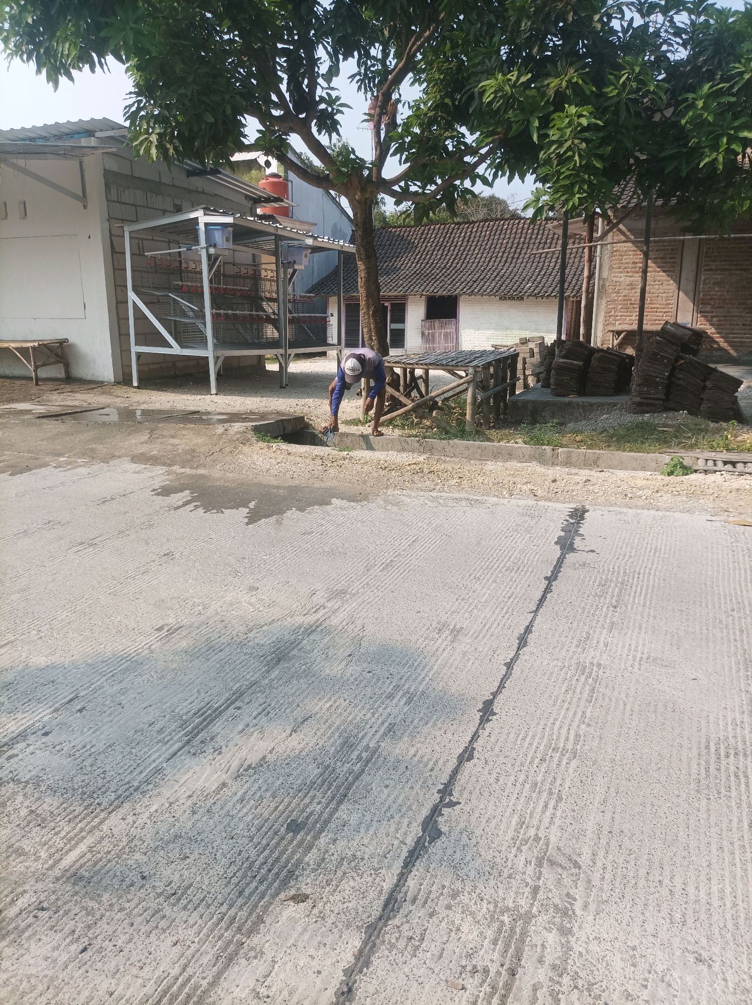 A man wearing a blue shirt and a white baseball cap is working near a drainage ditch in Canga'an, Indonesia. He is bent over, holding a tool or wire, and appears to be clearing debris from the ditch. The ground is a concrete surface with a rough texture, and there are some puddles of water present. In the background, there are several buildings with tiled roofs, a large tree with green leaves, and stacks of dark-colored tiles. A metal structure with multiple cages is also visible. The overall scene suggests an outdoor work area in a semi-rural or residential setting.