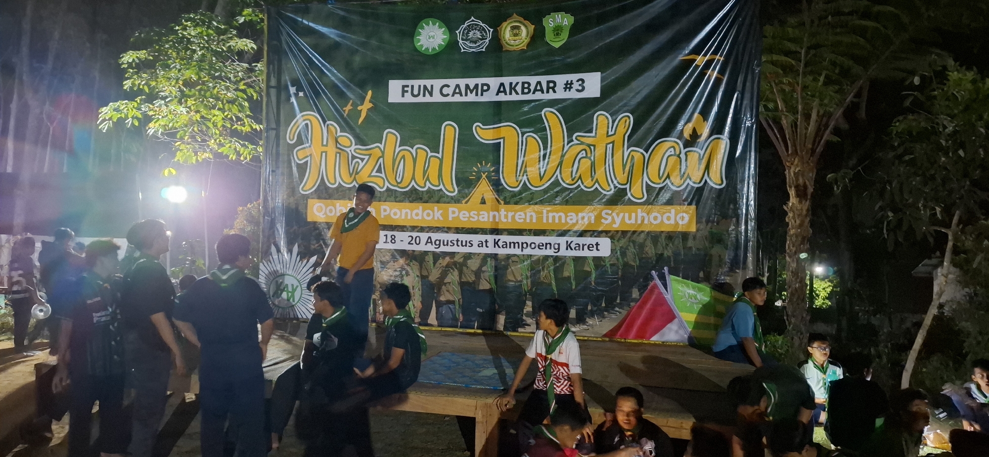 The image depicts an outdoor event at night, likely a camp or gathering, with a large banner in the background. The banner is dark green with yellow text and logos, advertising "FUN CAMP AKBAR #3 Hizbul Wathan" and mentioning "Pondok Pesantren Imam Syuhodo" and "18 - 20 Agustus at Kampoeng Karet." Several individuals, mostly young males, are visible. Some are standing, while others are seated on wooden platforms or the ground. Many are wearing scout-like uniforms or green neckerchiefs. The environment appears to be forested or park-like, with trees and foliage visible, illuminated by artificial lighting. The overall atmosphere suggests a community event. The location is Puntukrejo, Indonesia.