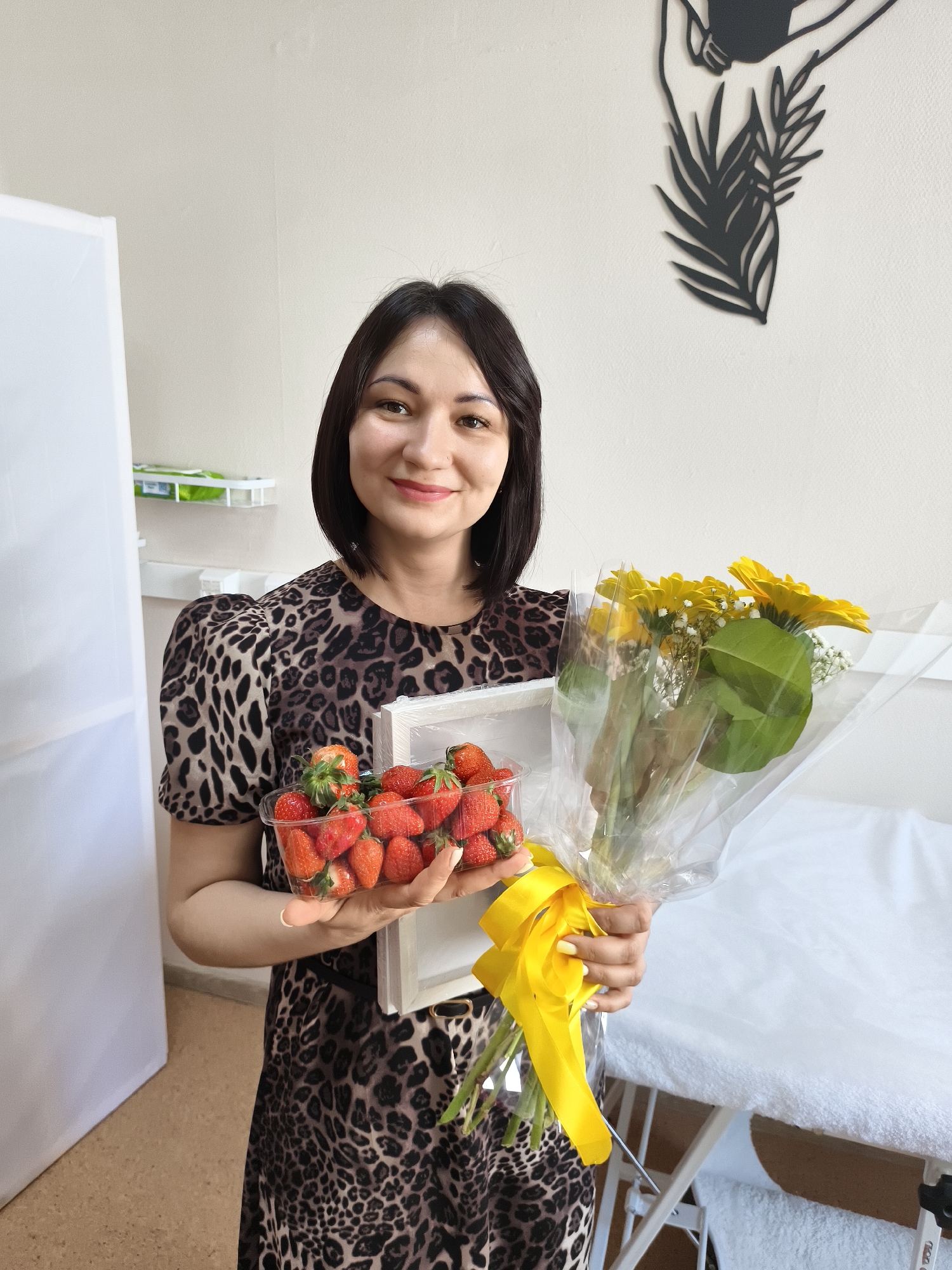 A woman is holding a plastic container of strawberries and a bouquet of yellow flowers wrapped in clear plastic with a yellow ribbon in what appears to be a medical or spa setting in the city of Unknown, Unknown. She has dark, shoulder-length hair and is smiling at the camera. She is wearing a leopard-print dress with short, puffed sleeves. In the background, there is a white room divider and a black metal wall decoration of leaves. To the right of the woman, there is a white examination table with a white towel on it. The floor is tan-colored.