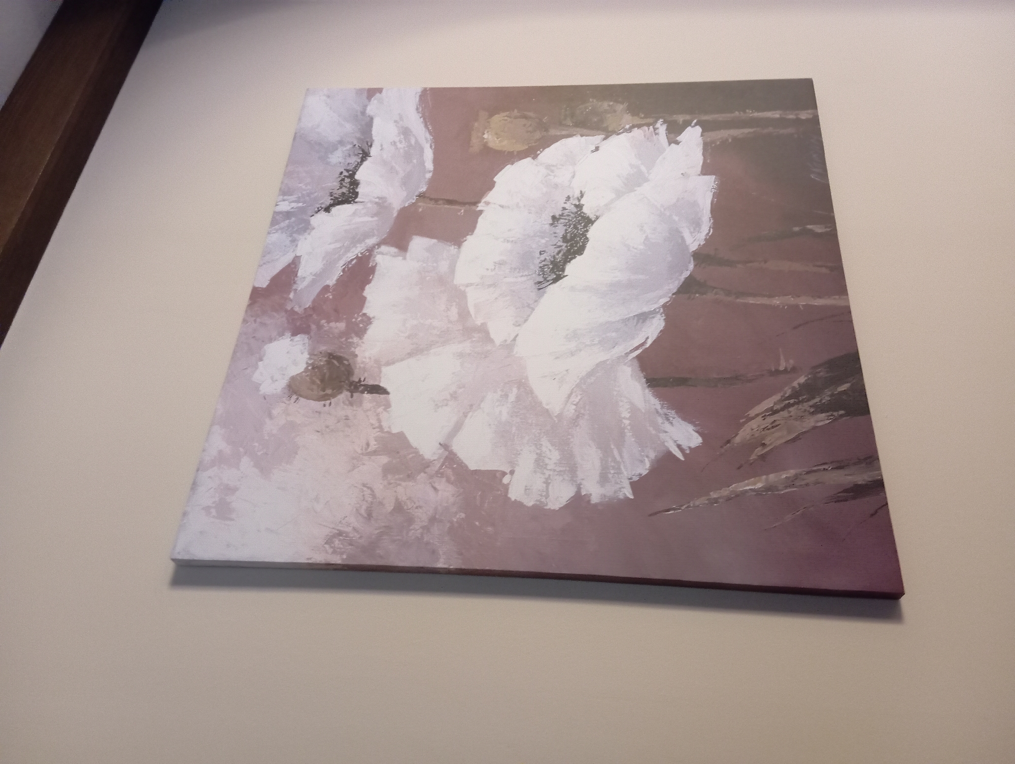 An abstract painting of white poppies hangs on a plain, beige wall. The painting is rectangular and oriented horizontally. The poppies are rendered with thick, brushstroke textures, predominantly in white and pale pink, against a muted, dusty rose background. Several tall, thin green stalks are visible in the foreground. A dark brown wooden beam runs horizontally across the top of the frame, casting a shadow on the wall above the painting. The painting is signed in the bottom right corner, though the signature is illegible. The scene is indoors, likely in a domestic or gallery setting. The location in Baden, Austria, is not specified by any visual cues in the image.