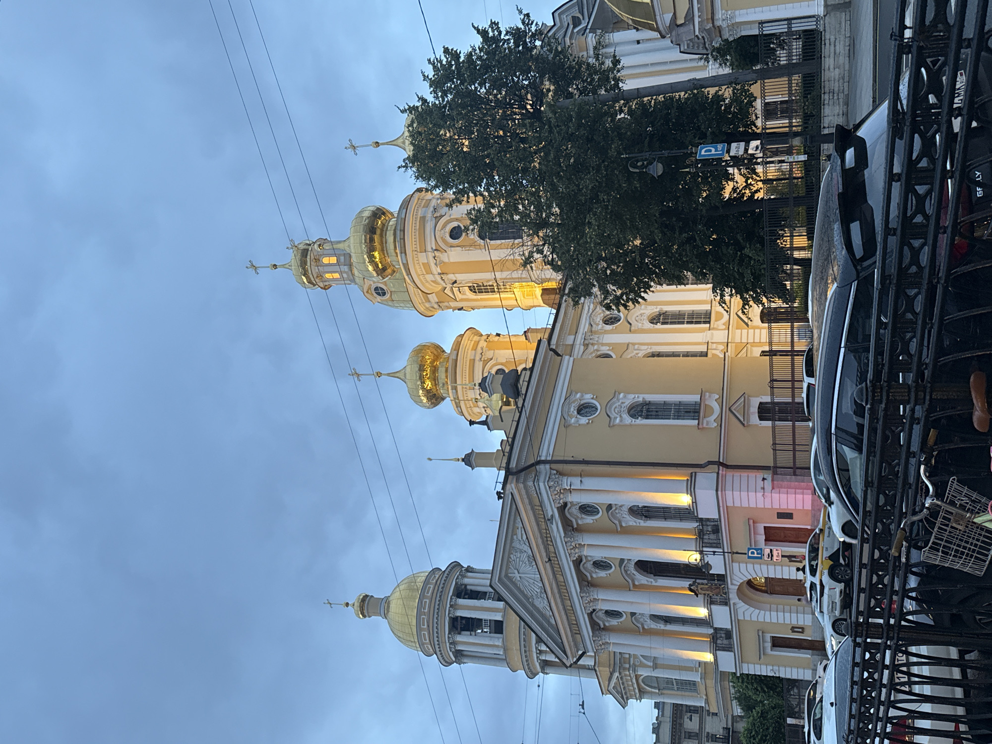 The image shows a yellow Orthodox church with multiple gold domes and crosses, illuminated by artificial lights. The church has a classical facade with columns and arched windows. A large green tree stands to the right of the church, partially obscuring it. In the foreground, a black metal fence runs horizontally across the bottom of the frame. Behind the fence, several cars are parked on a street, including a white sedan and a yellow taxi. The sky is overcast and a pale blue. Power lines are visible against the sky.