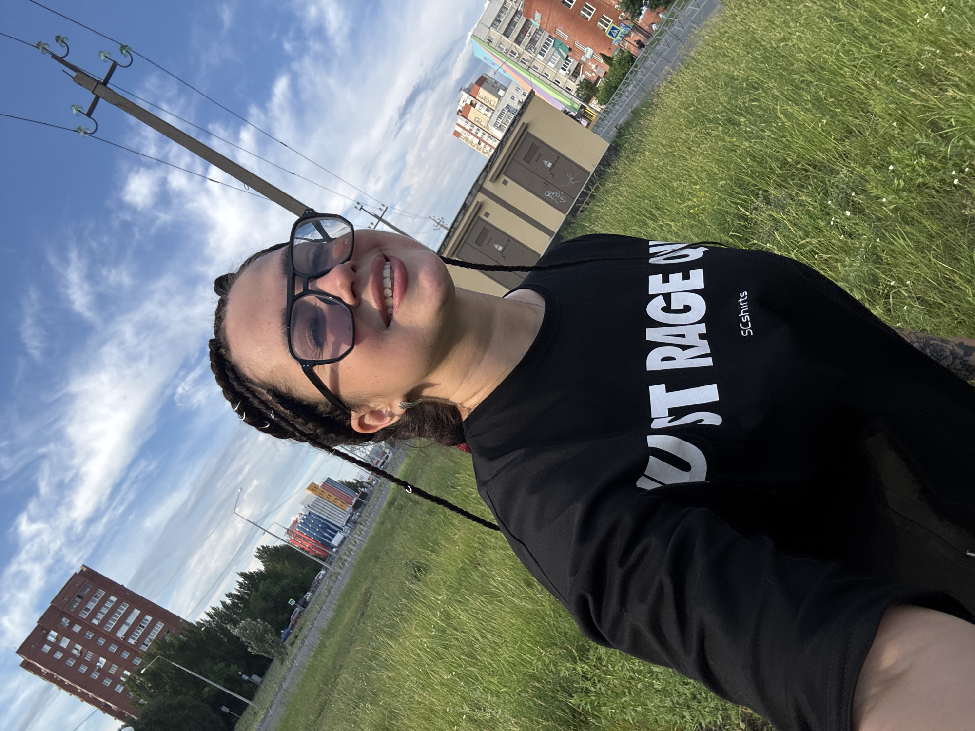 A young woman with dark braided hair and glasses smiles at the camera in a selfie taken outdoors. She is wearing a black long-sleeved shirt with white text that reads "RAGE ON" and "SCShirts". The background features a grassy field, a street with cars, and several multi-story buildings. A utility pole with wires is also visible. The sky is blue with scattered clouds.
