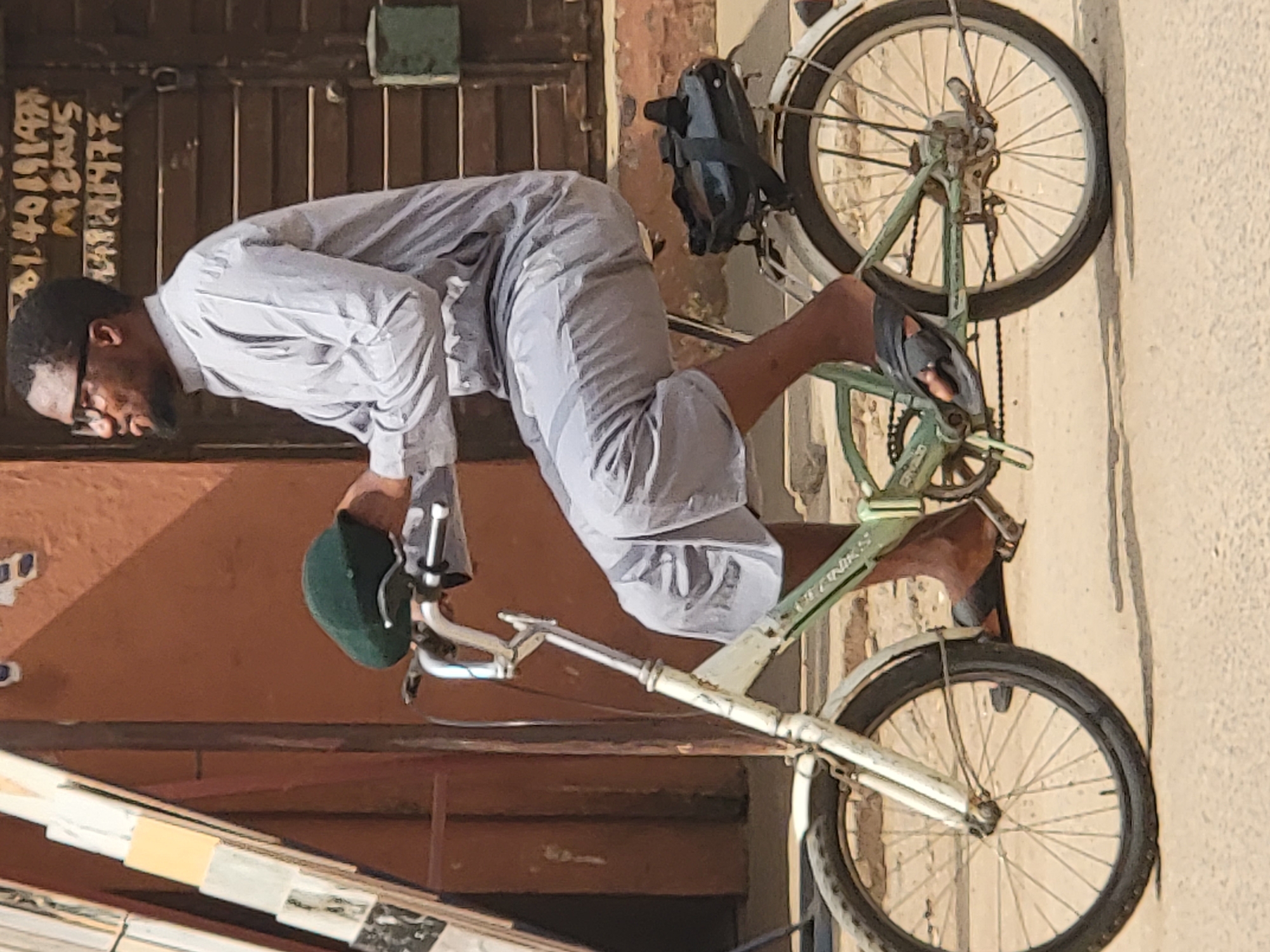 A man is seen riding a bicycle in what is likely a street of Maiburiji, Nigeria. The man has dark hair, a beard, and is wearing glasses. He is dressed in a light-colored, long-sleeved shirt and trousers. He is holding a green cap in his left hand. The bicycle is old, with a green frame and rusted white handle bars. A black backpack is attached to the bicycle. The location is suggested by writing on the background wall. The street appears to be outdoors with buildings in the background. The man seems to be actively riding the bicycle.