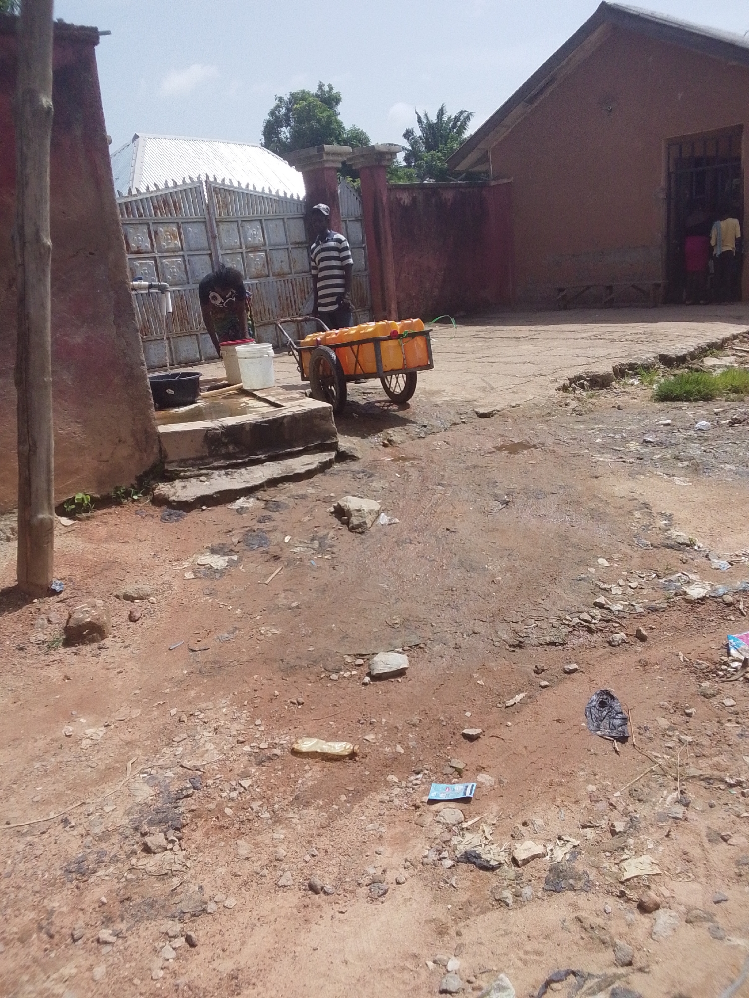 The image depicts an outdoor scene in what appears to be a rural or semi-urban area, possibly Jalingo, Nigeria. In the foreground, a dusty, unpaved ground is visible, littered with rocks, debris, and some scattered plastic waste, including a plastic bottle and a black plastic bag.
The main subjects are two individuals and a handcart loaded with yellow plastic containers, likely filled with water. One person, wearing a striped shirt and a cap, is standing near the handcart. Another person, wearing a dark t-shirt with a graphic design, is bent over near a water source, which consists of a concrete structure with a tap and a black basin. Two buckets, one white and one red, are also present near the water source.
Behind the individuals and the handcart, a large metal gate with a decorative pattern is visible. Beyond the gate, a building with a white, corrugated metal roof stands out. To the right of the gate, a pinkish-brown building with a dark, barred entrance is partially visible.
The activity taking place appears to be the collection or distribution of water, given the presence of the handcart laden with containers and the water source. The lighting suggests it is daytime, with a bright, overcast sky. The overall impression is one of a functional, perhaps everyday scene related to water access in the community. There is no visible text in the image.