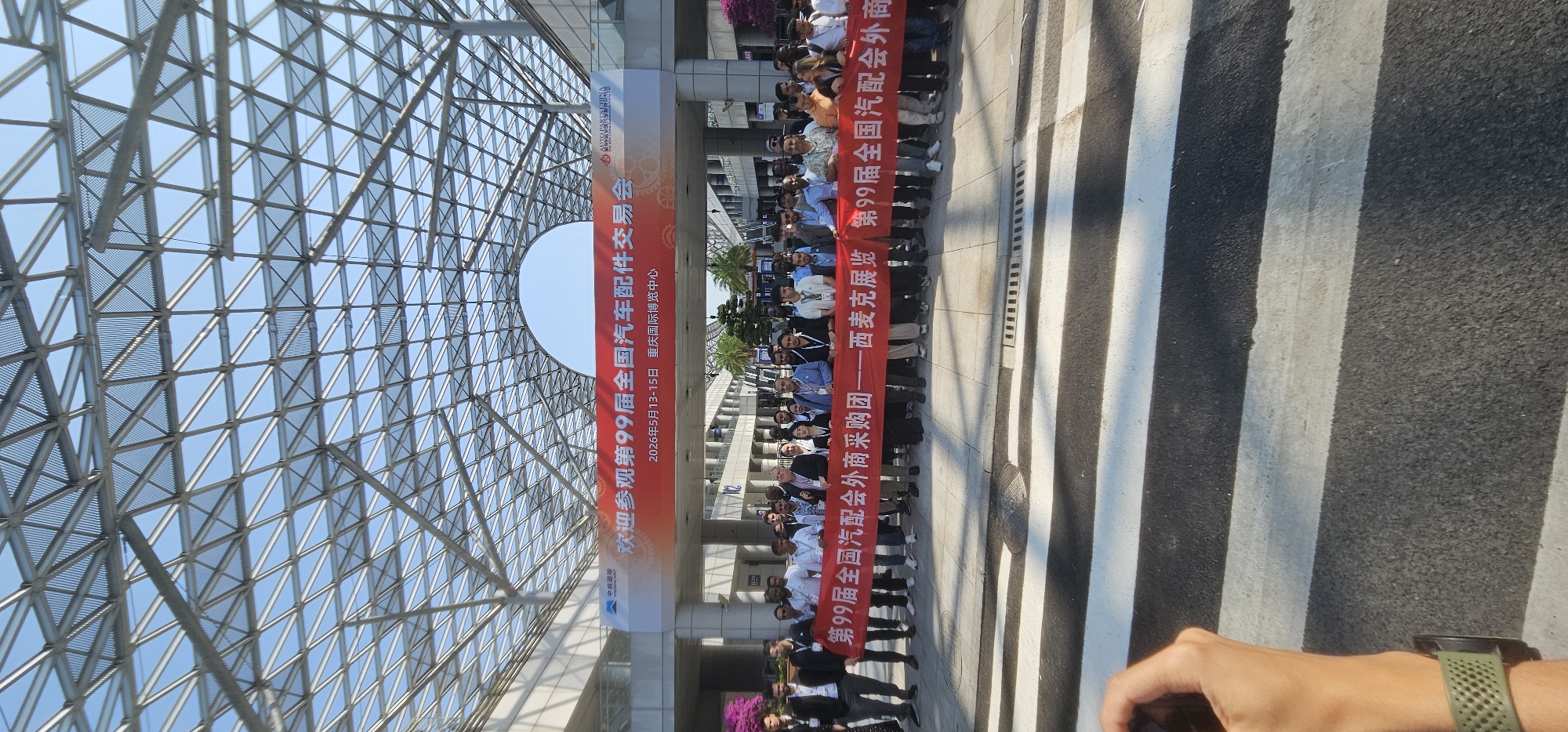 A wide-angle photograph shows a group of people holding a banner outside the Liangjiang International Expo Center in Chongqing, China. The banner reads in Chinese: "Welcome to the 99th National Auto Parts Fair." The banner also includes the dates of the fair, May 13-15, 2026. The group of people appear to be attending or participating in the event. Above the group is a large, arched glass and steel structure that covers the entrance to the expo center. The location is outdoors, and the time of day appears to be mid-morning or early afternoon. The camera is tilted slightly.