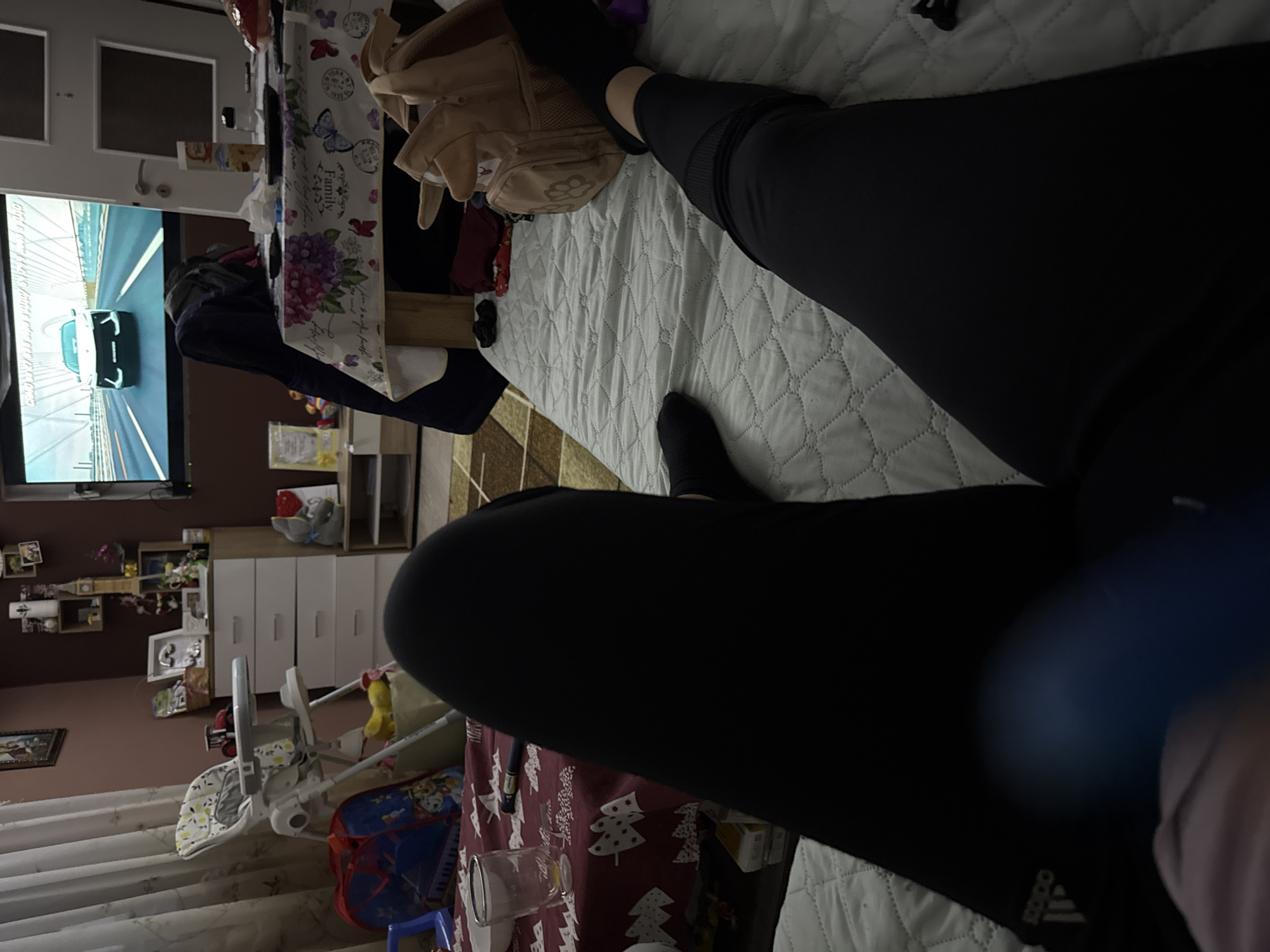 The image shows a person's legs wearing black leggings lying on a light-colored quilted surface. A TV screen showing a racing game is visible in the background. A table with a floral-patterned cloth is beside the person. A baby high chair and a box with a Christmas-themed cloth are present in the room. Other furniture and decorations are also visible, but the exact location in Sofia, Bulgaria, is not discernible from the image alone.
