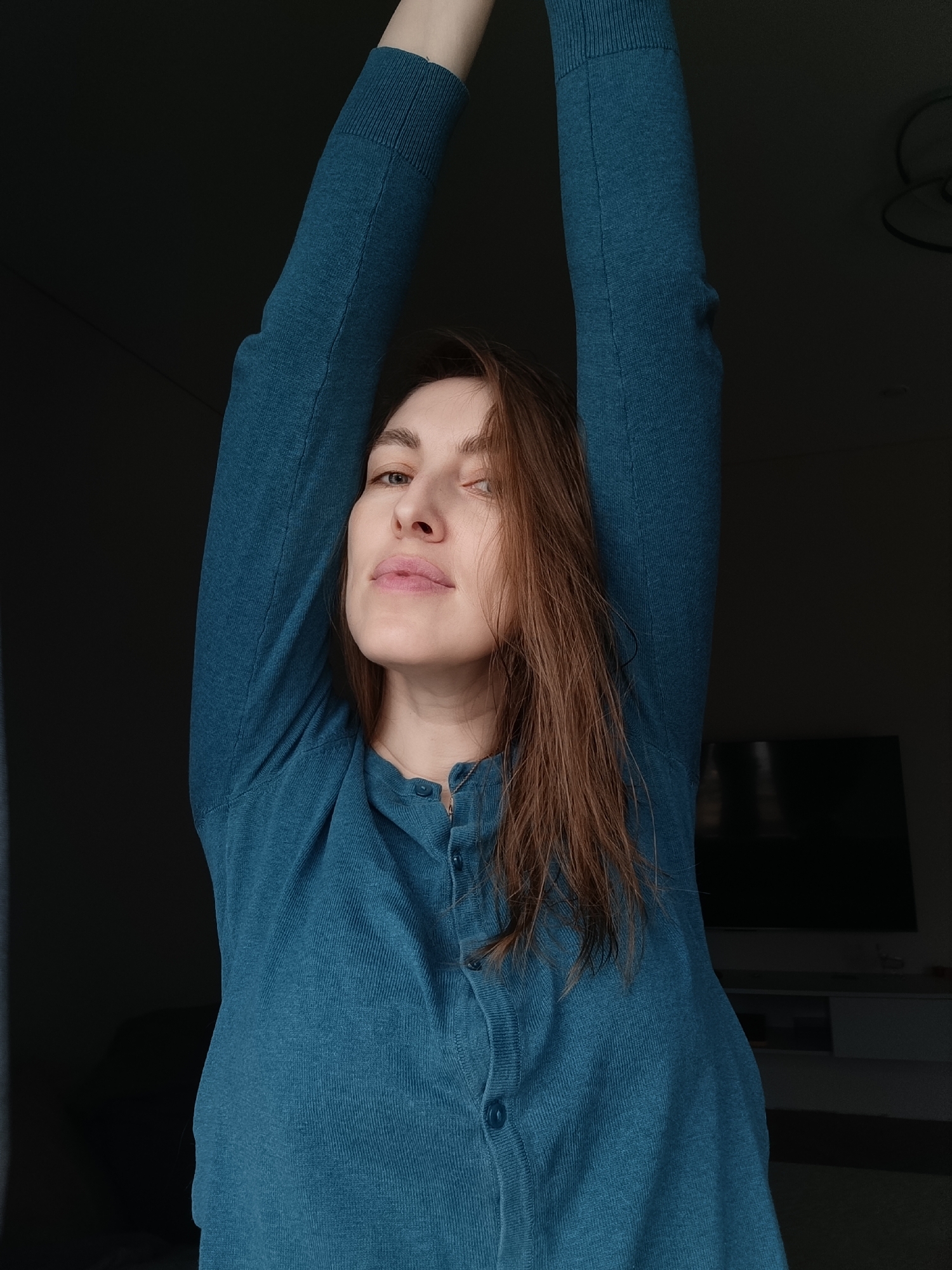 A woman with shoulder-length brown hair is the central subject of the image, wearing a long-sleeved, blue-green button-down cardigan. Her arms are raised above her head, suggesting a stretching or reaching gesture. Her face is visible, and she is looking towards the camera.
The background is dimly lit, and a television set is discernible on a white cabinet. The location is reported as Chaikovsky, Russia, but there are no visual cues in the image to confirm this.