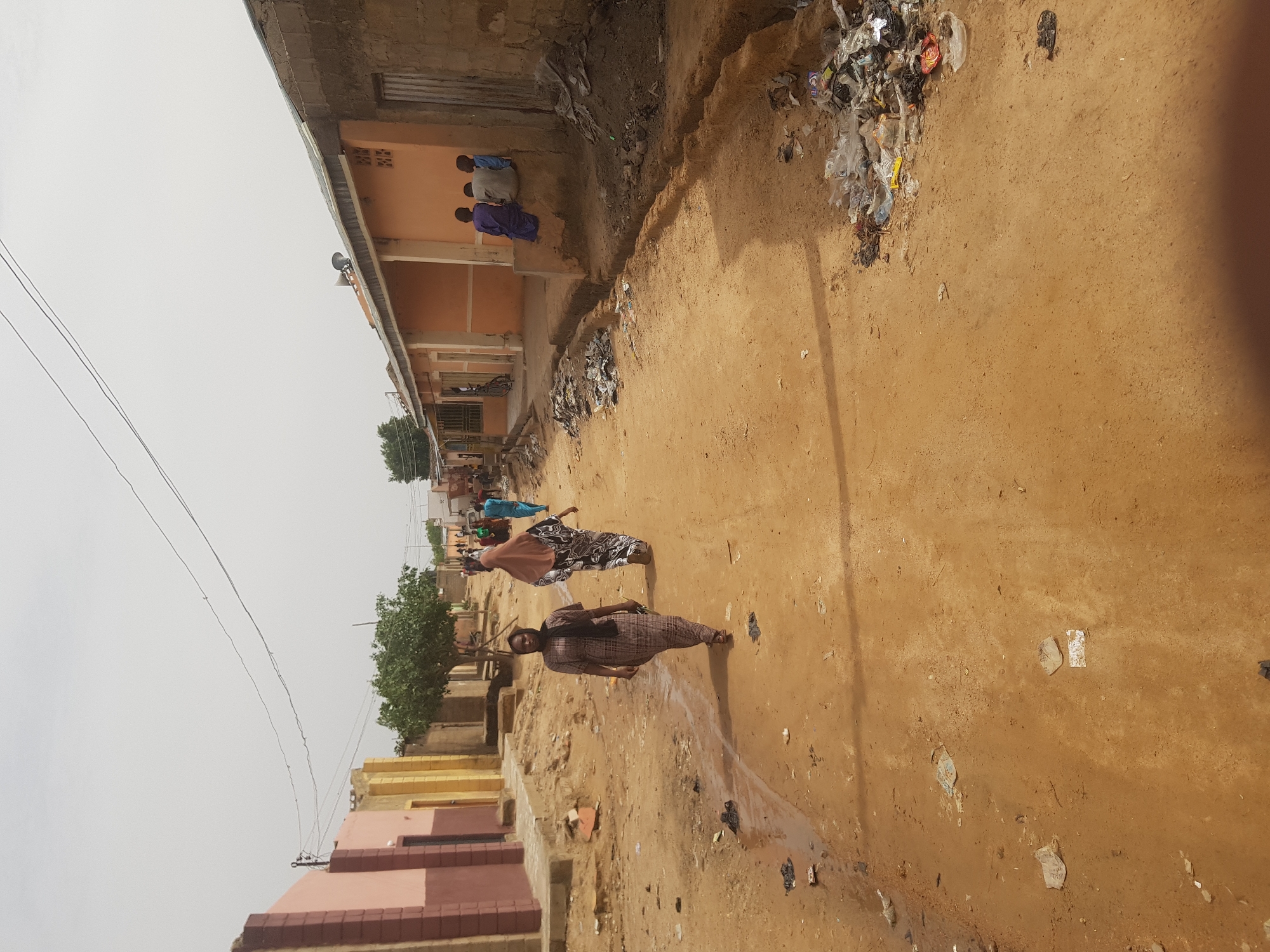 The image depicts a street scene in Dabawa, Nigeria. The road is unpaved and covered in dirt. Litter and debris are scattered along the roadside and in a large pile near a building on the right. Several people are walking along the road. One woman in the foreground is wearing a patterned dress and a headscarf. Another woman further down the road is wearing a light brown shawl. In the background, there is a group of people wearing blue and purple clothes on a balcony. Buildings line the street. Some are constructed of brick or concrete. Power lines stretch across the sky. The sky is overcast and pale.