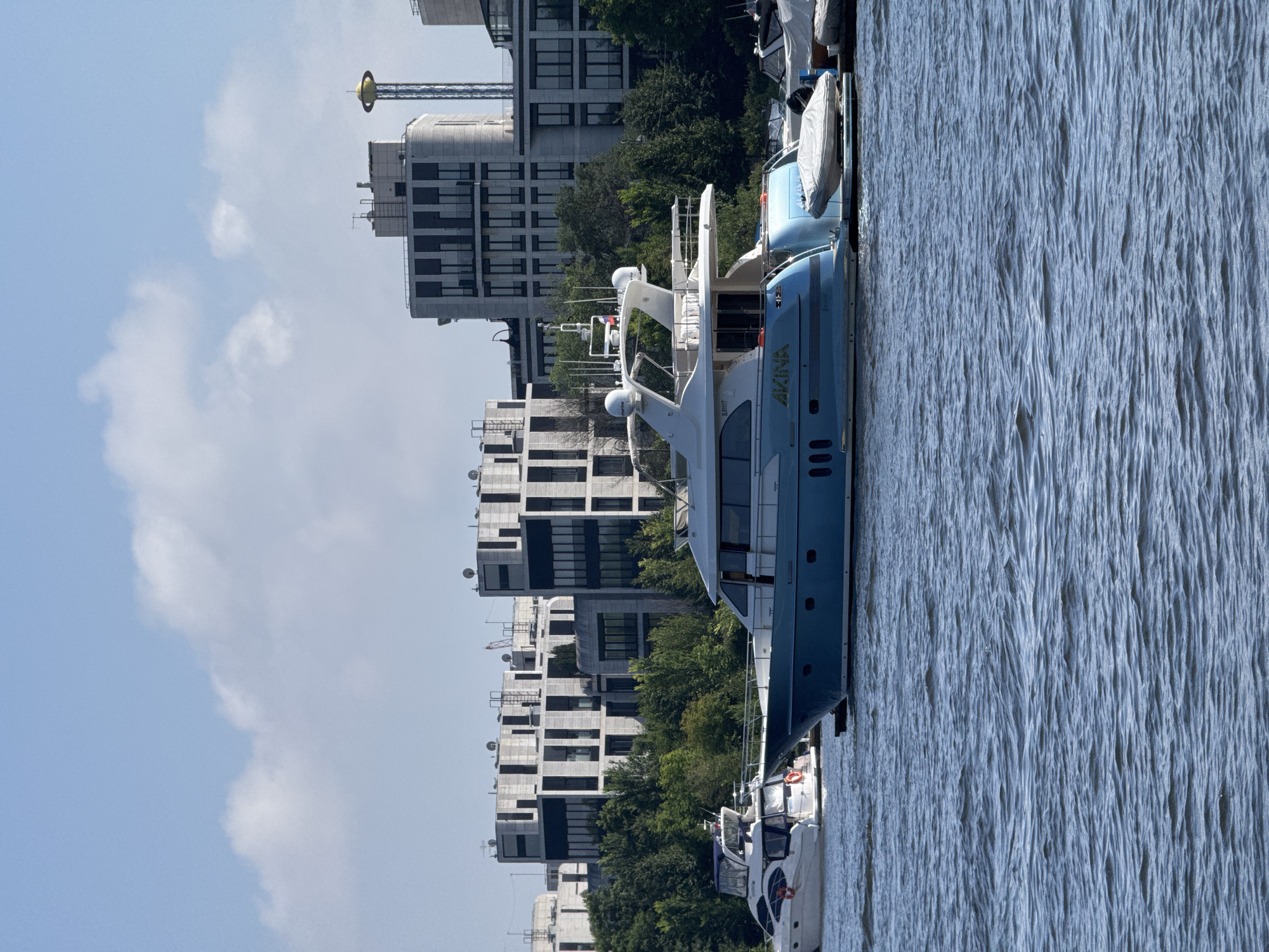 This media file captures a view of a blue yacht named "AKINA" docked in a body of water, likely a river or canal, with buildings and trees on the shore. The buildings are modern, with many windows and balconies, and are interspersed with lush green trees, suggesting an urban environment. The water is calm with visible ripples. The sky is a clear blue with a few white clouds. The image appears to be taken on a bright, sunny day, likely in the afternoon. The overall impression is one of a peaceful urban waterscape. The yacht is prominently featured, with its name clearly visible on the hull. Other smaller boats are also docked alongside. The scene suggests a leisurely setting, possibly a marina or waterfront promenade.
