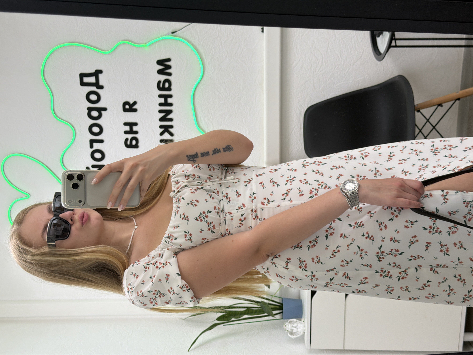 A young blonde woman with long hair, wearing sunglasses, a white floral dress, and a beaded necklace, takes a mirror selfie in a room. She is holding a smartphone in her right hand. The wall behind her has a green neon sign with black text. A plant in a blue pot sits on a white cabinet to her left. A black chair and a small black table are visible in the reflection of the mirror. The scene appears to be an interior space, possibly a shop or a dressing room. The neon sign's text and the overall ambiance suggest a location in a city, and the reference to Chaikovsky, Russia, indicates a specific geographical context.