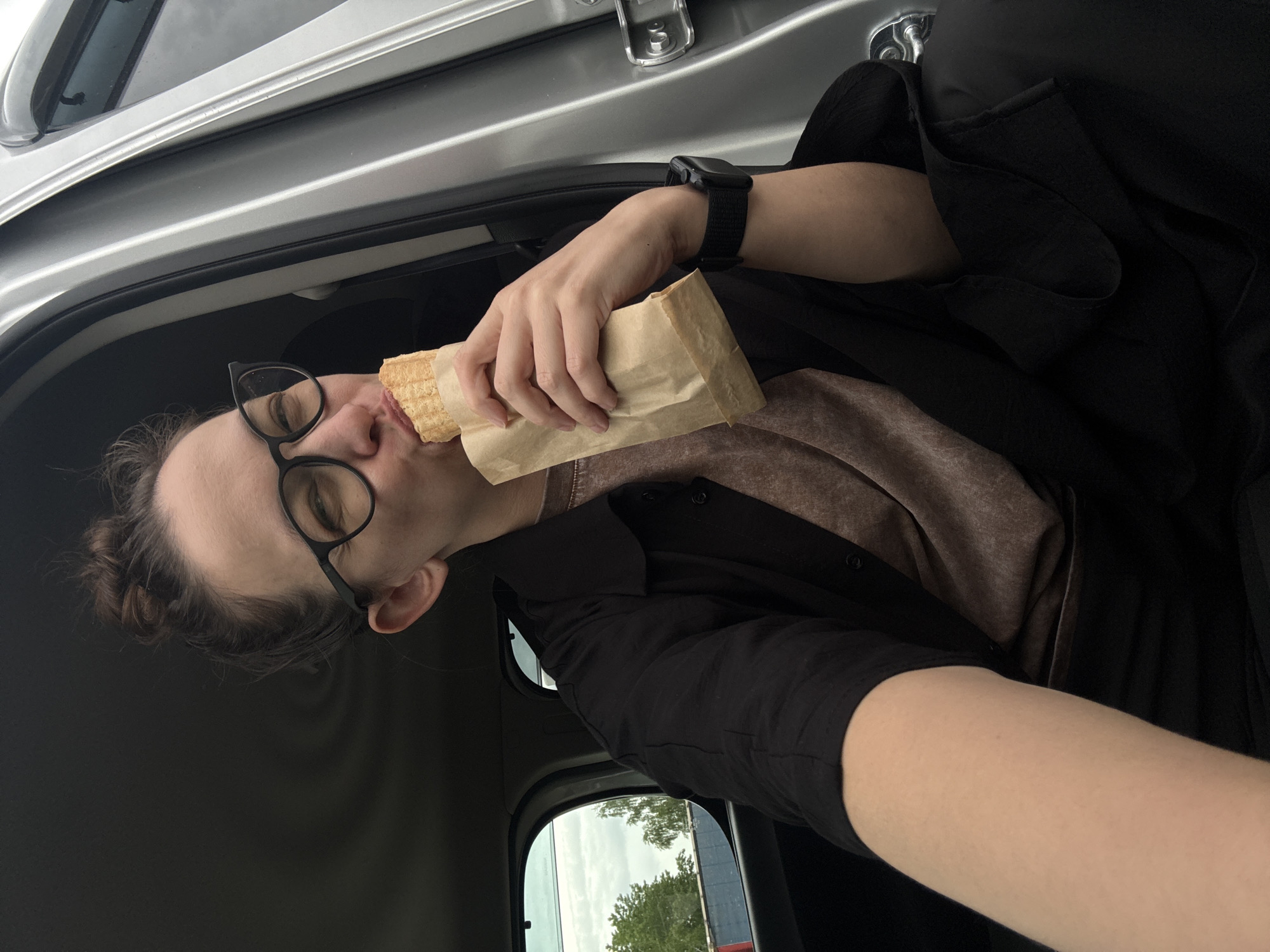 A person with glasses and a bun is eating from a paper bag inside a vehicle. The person is wearing a black watch, a black shirt over a brown shirt, and is positioned to the right of the frame, holding a paper bag to their mouth. The vehicle's interior is visible, including a window with a view of trees and a building. Based on the provided information, the location is labeled as Unknown, Russia.