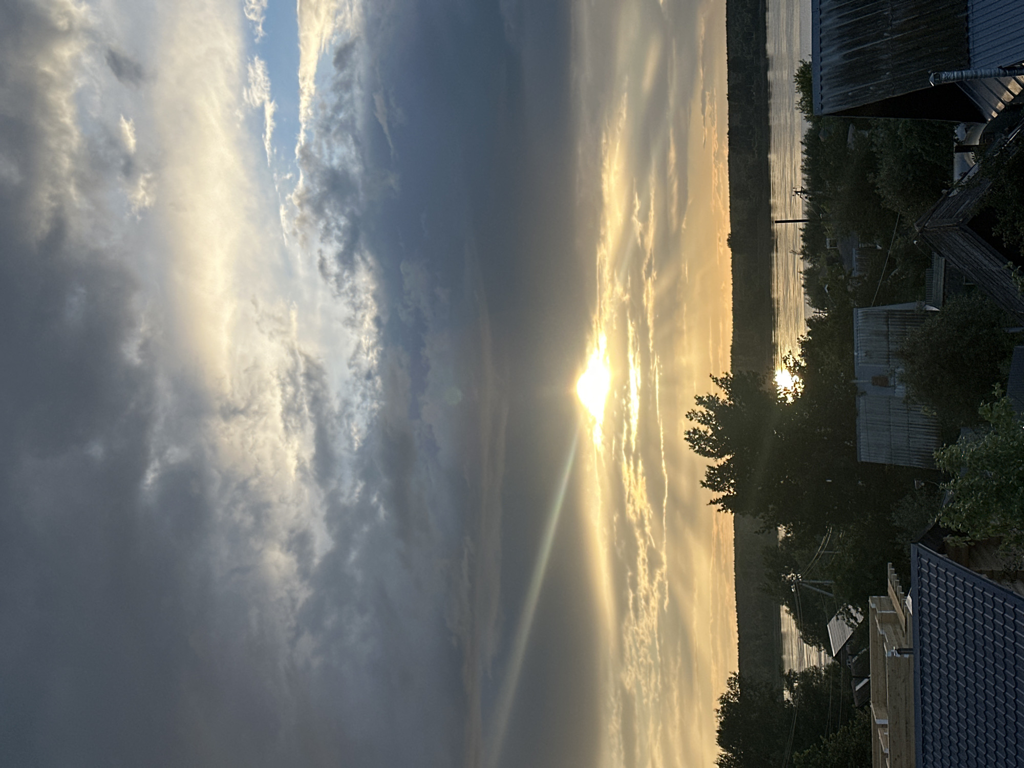 The image depicts a sunset over a body of water, likely a river or lake, in Chaikovsky, Russia. The sky is filled with dramatic clouds, transitioning from dark, stormy grey on the left to bright, golden hues where the sun is setting on the right. Sunbeams pierce through the clouds, creating a luminous effect. In the foreground, the rooftops of several houses are visible, some appearing to be under construction with exposed wooden beams. Dense trees and foliage surround the houses and line the distant shore. The water reflects the warm colors of the sunset, creating a shimmering effect. The overall mood is serene and picturesque, characteristic of a late summer evening. There is no visible text in the image. The time of day is clearly sunset, and the weather appears to be clearing after some cloudiness, allowing for a vibrant display of natural light.
