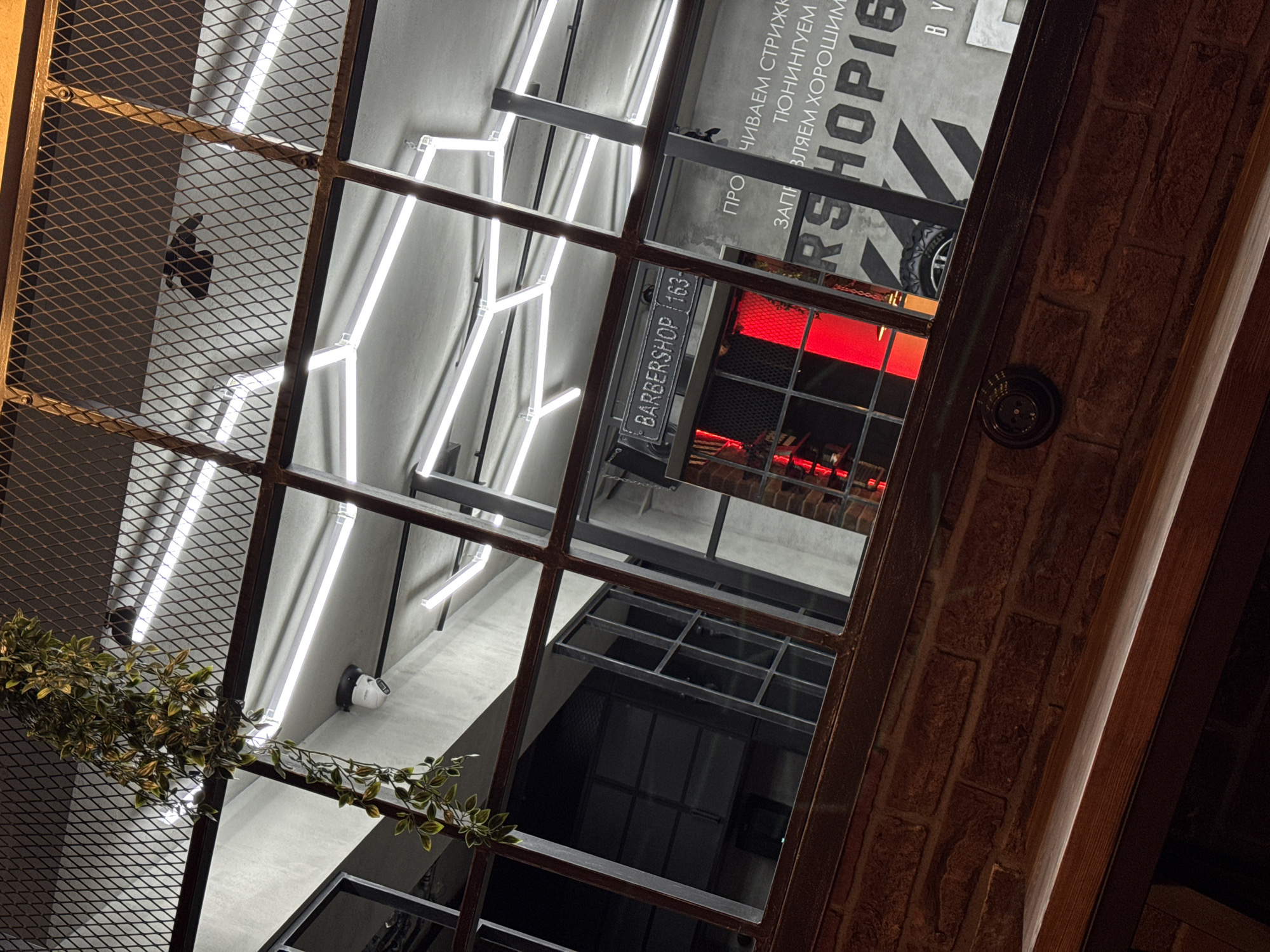 The image captures an interior view through a window or a framed opening, possibly within a building in Tolyatti, Russia. The view features elements of an establishment, which appears to be a barbershop. This is indicated by the signage "BARBERSHOP 163" visible within the frame. The interior design includes a modern lighting arrangement of fluorescent tubes forming abstract patterns on the ceiling. Parts of a brick wall are visible around the frame. The environment appears contemporary, with a mix of industrial and modern decorative elements.