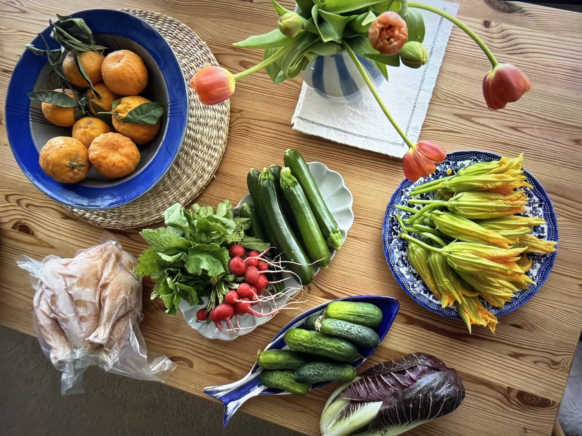 An overhead view captures a vibrant array of fresh produce and flowers arranged on a light wooden table.
In the upper left, a blue bowl holds several oranges, some still bearing green leaves, resting on a round woven straw placemat. Below this, a whole raw chicken, still in its clear plastic packaging, is visible. Adjacent to the chicken, a bunch of red radishes with their green tops is placed next to multiple green zucchinis in a white scalloped dish.
Further down on the table, a distinctive blue and white fish-shaped dish contains several small cucumbers. To its right, a head of purple radicchio or red cabbage lies on the wooden surface.
In the upper center of the frame, a blue and white striped vase holds a bouquet of green-stemmed tulips. Some tulips are open, displaying orange-red petals, while others remain in bud. The vase sits on a folded white cloth. To the right of the flowers, a blue and white patterned plate is filled with numerous pale yellow-orange squash blossoms.
The scene depicts a rich collection of fresh ingredients, suggesting preparation for a meal. The lighting indicates daytime. There are no discernible location cues or visible text within the image.